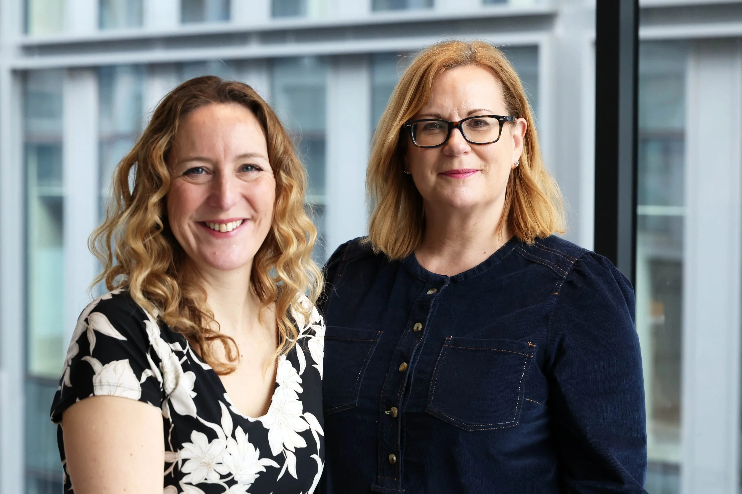 Two women standing indoors in front of large windows, smiling at the camera. The woman on left has curly blonde hair and is wearing a black and white floral dress. The woman on the right has blonde hair and glasses, and is wearing dark denim dress.