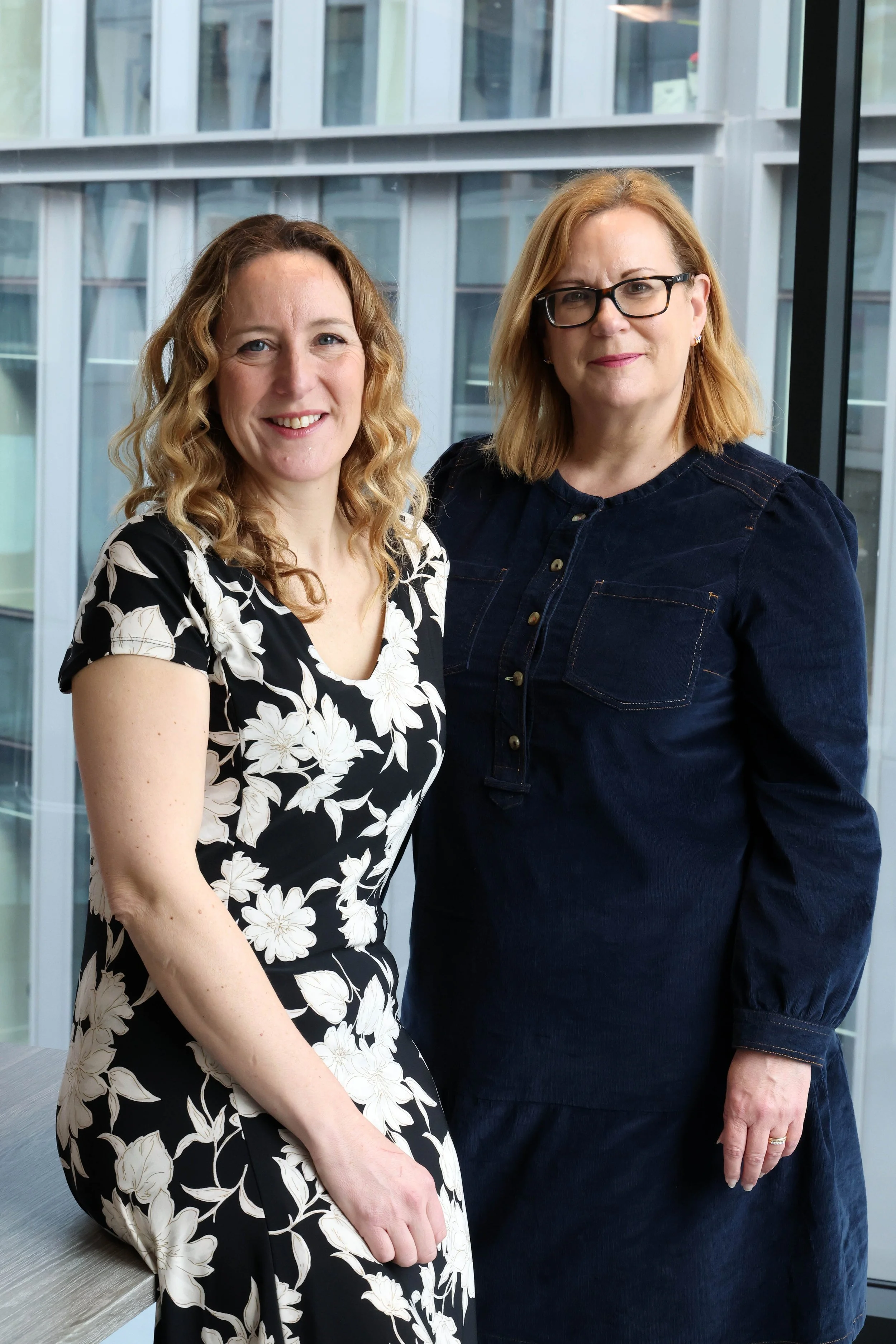 Two women, one with curly blonde hair wearing a black and white floral dress, and the other with straight blonde hair and glasses wearing a dark denim dress, standing together in front of large windows with a modern building exterior in the background.