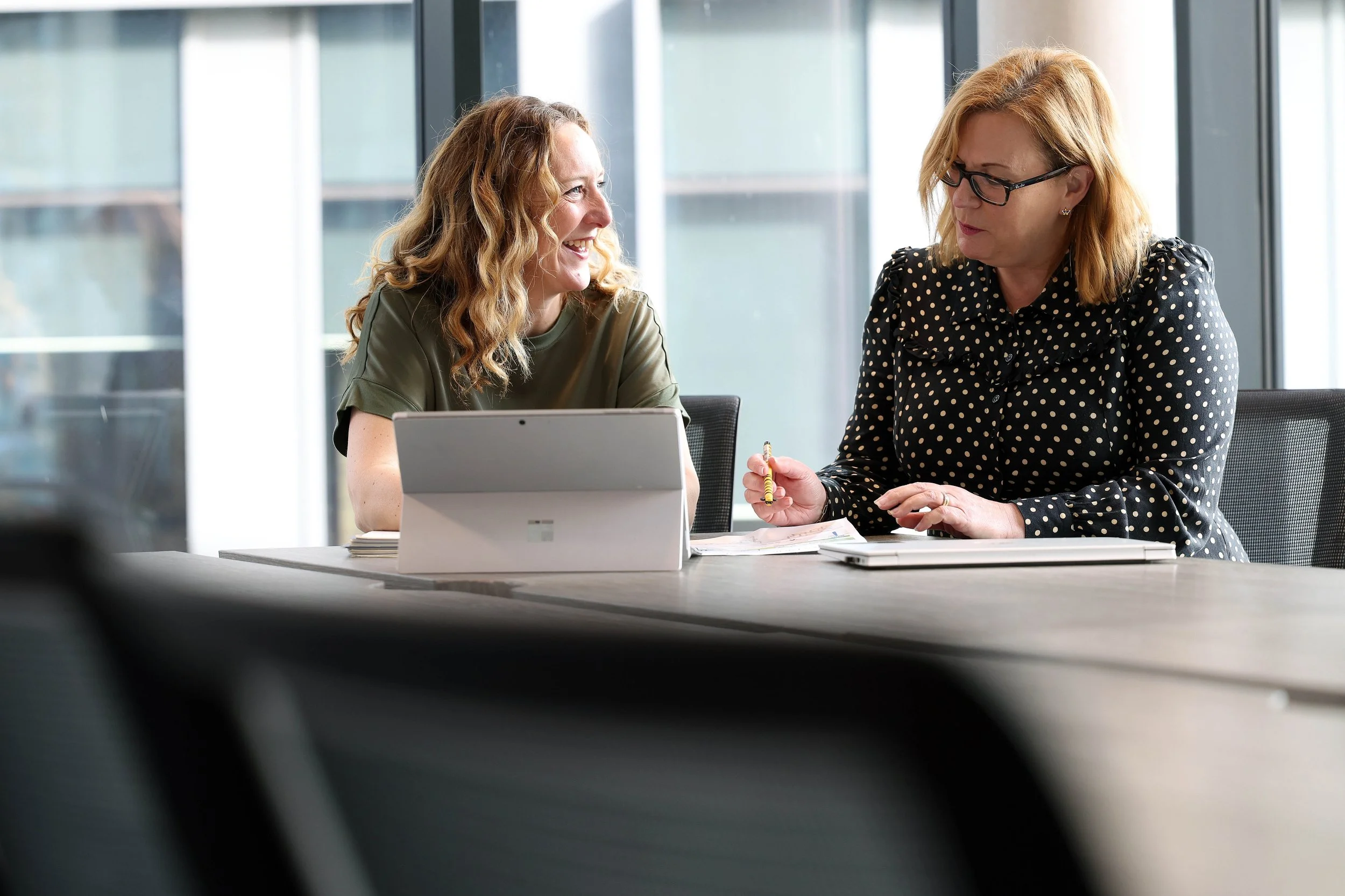 Two women sitting at a desk in an office, looking at a computer screen, with one woman pointing at it with a pen.