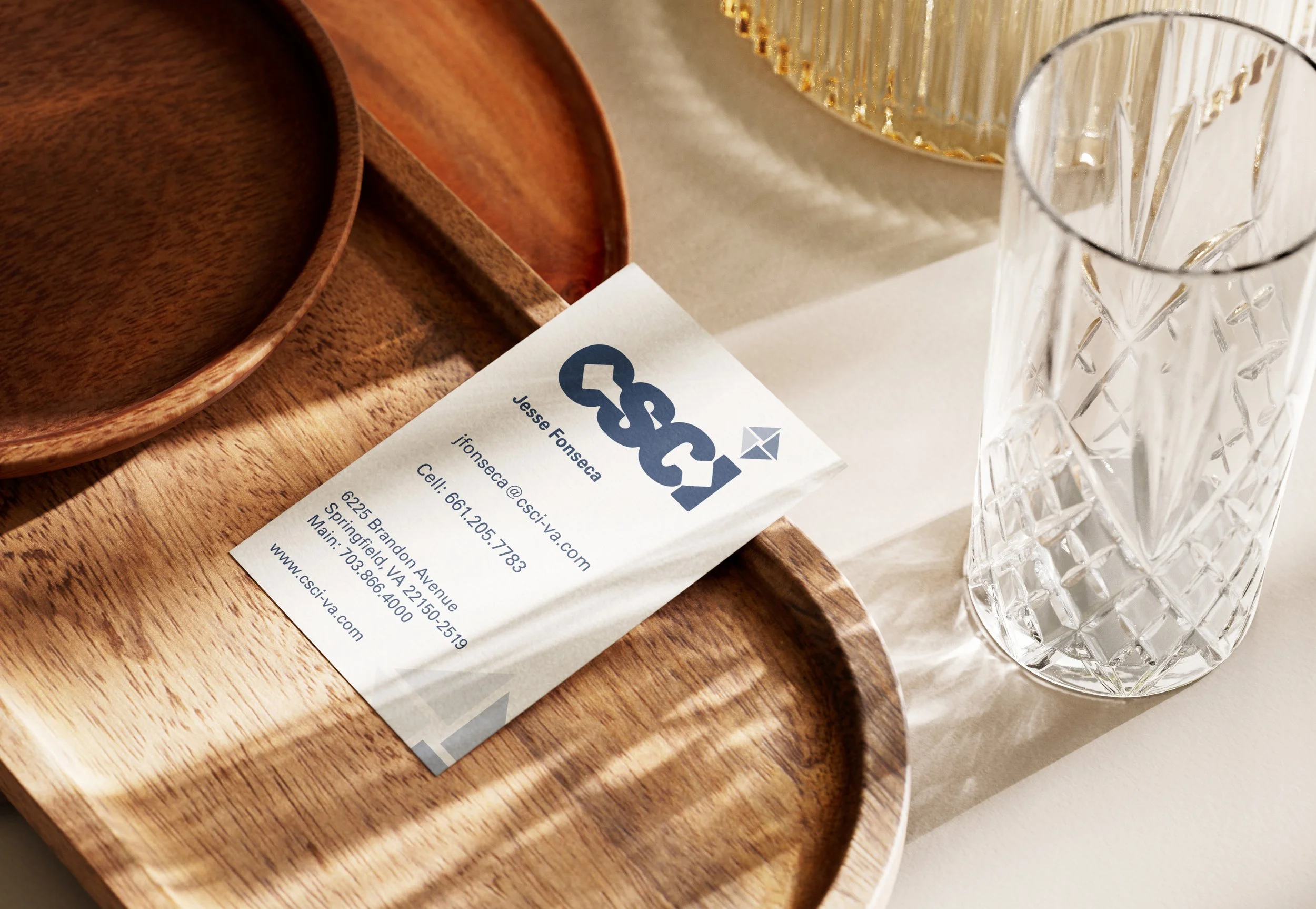 Business card for Jesse Fonseca placed on a wooden tray next to a clear glass with cut pattern, with a gold decorative item in the background.