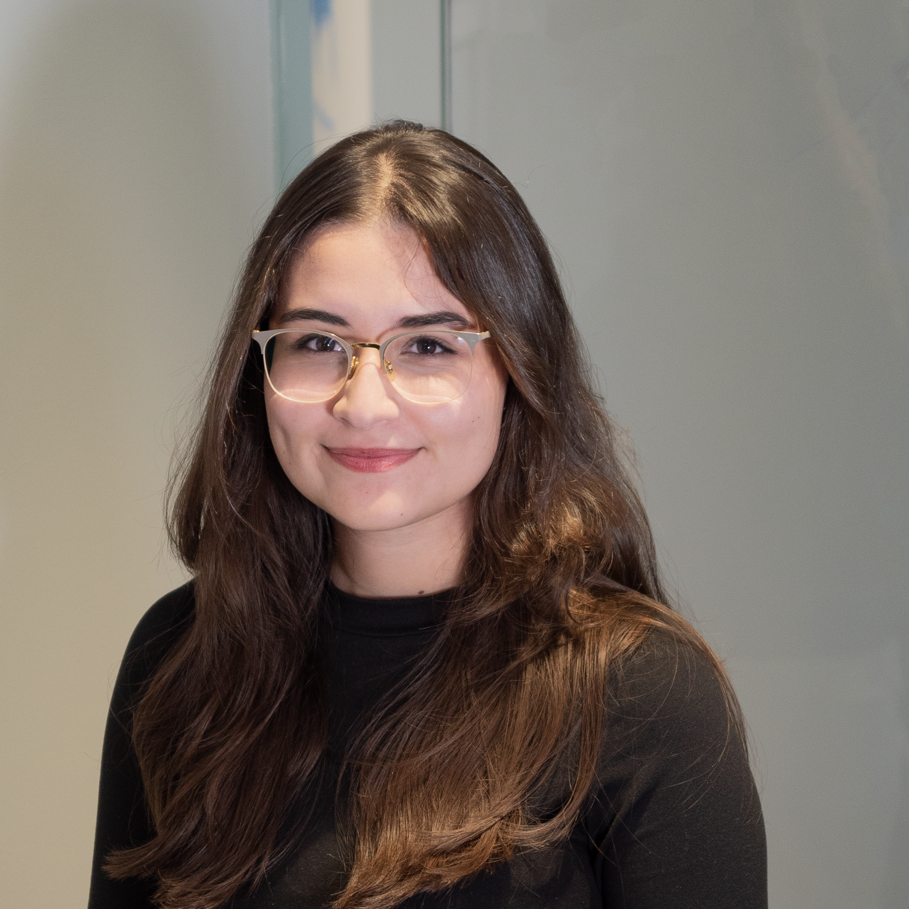 A young woman with long brown hair, glasses, and a black top, smiling at the camera indoors.