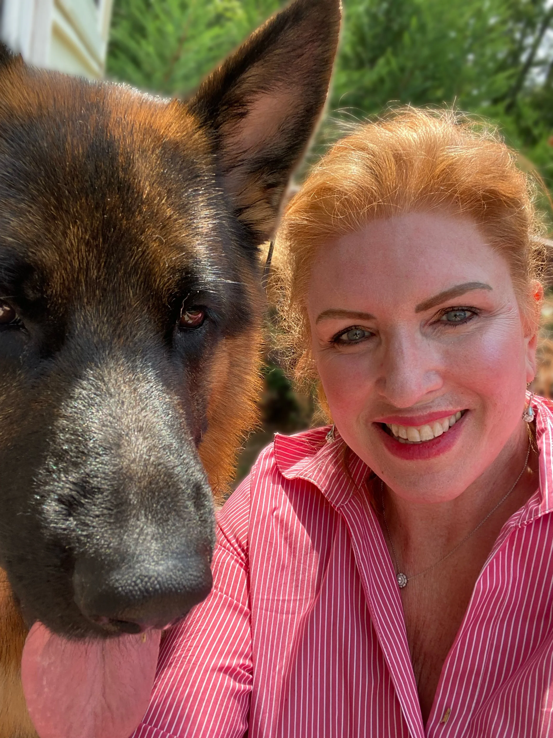 A woman with red hair, wearing a pink striped shirt, smiling next to a large brown and black dog with an expressive face and big ears, outdoors with greenery in the background.