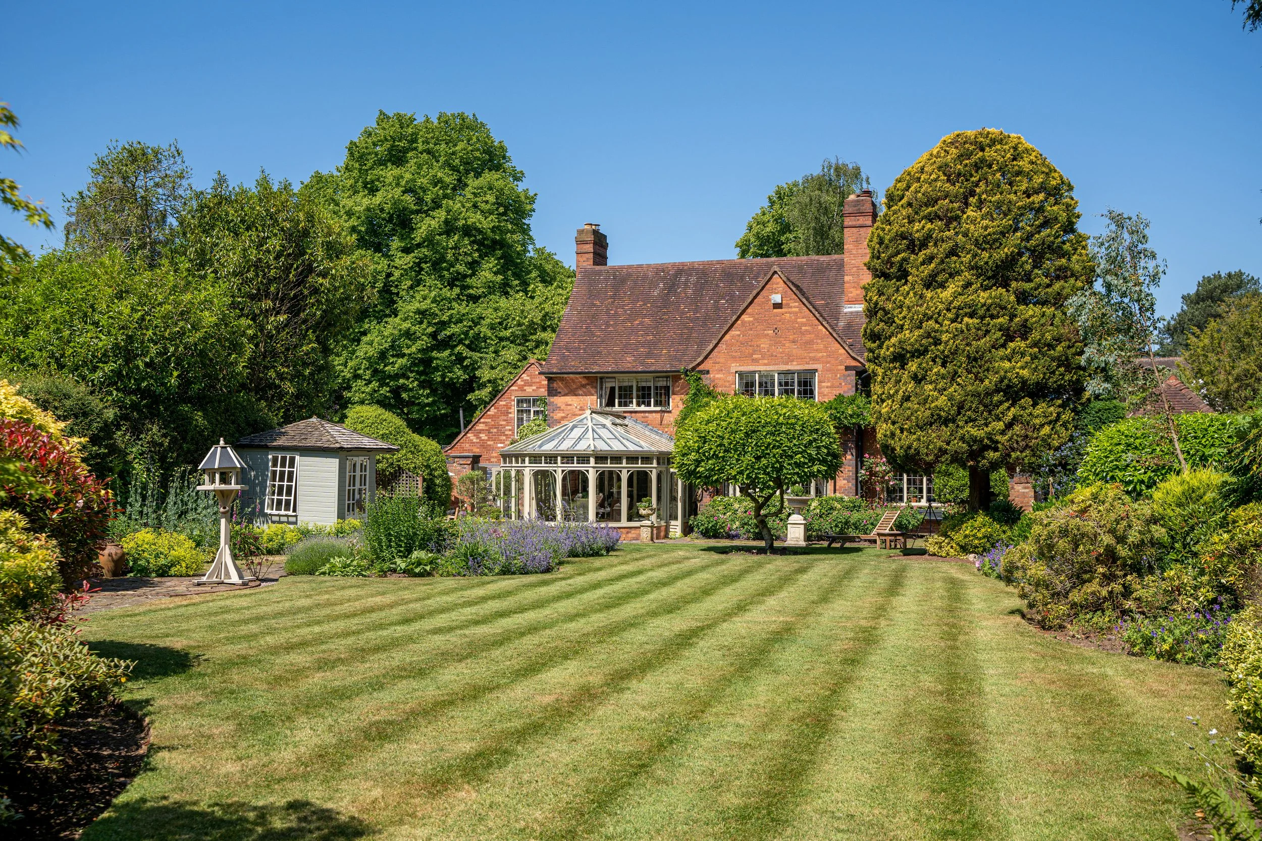 A spacious, well-maintained backyard with a large, striped lawn leading to a brick house with a conservatory and lush greenery, including trees and flower beds, under a clear blue sky.