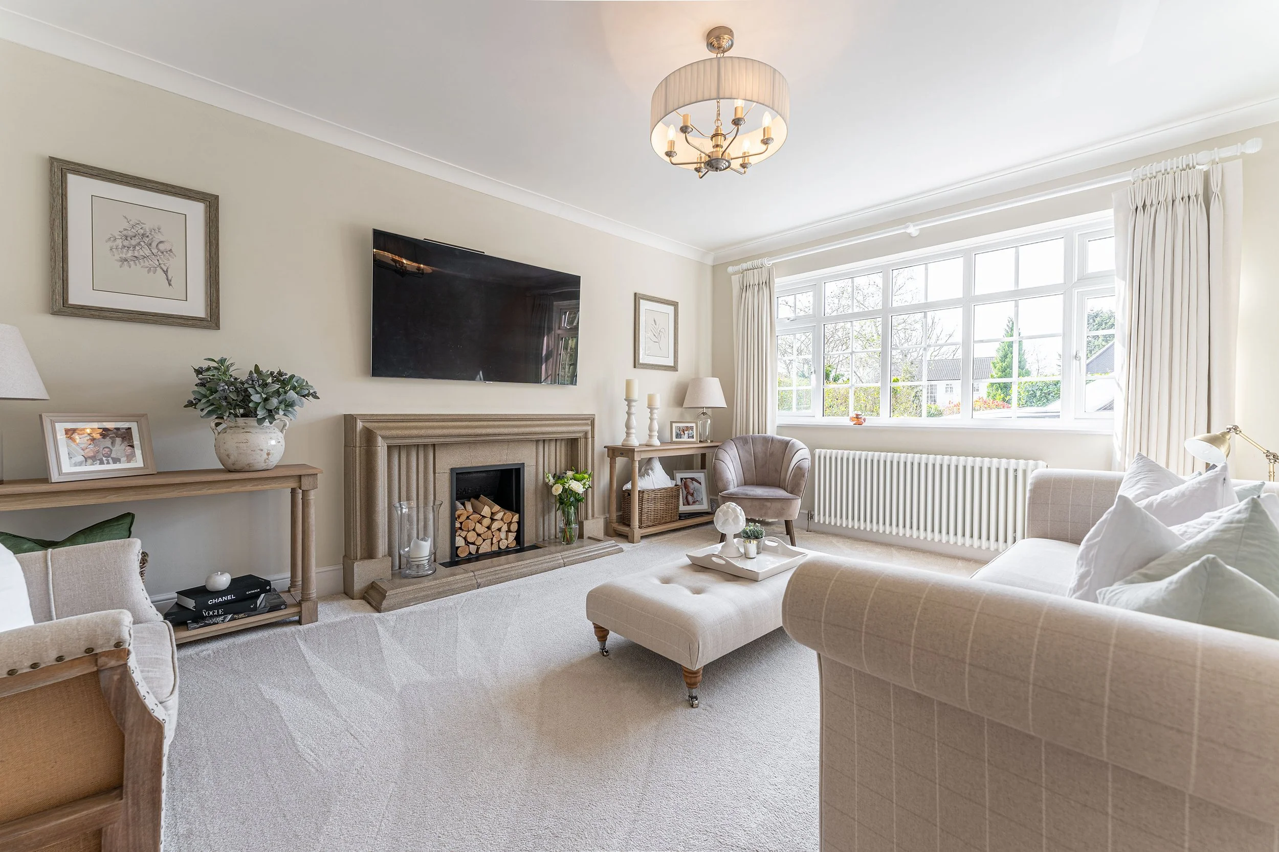Bright living room with large window, beige sofa, armchair, fireplace, wooden furniture, wall-mounted TV, neutral decor.