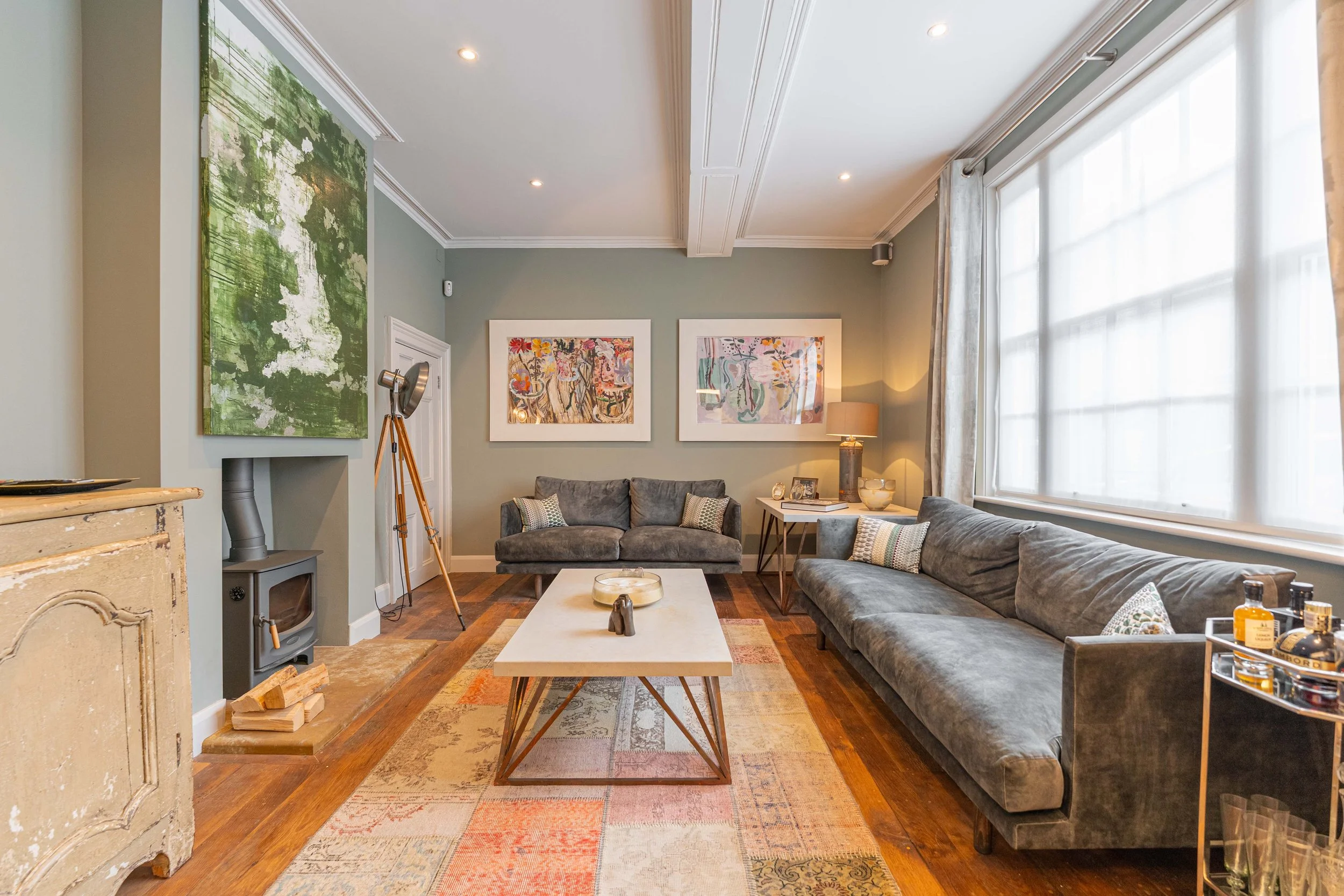 Living room with gray sofas, colorful artwork, a large window with curtains, a vintage fireplace, a coffee table, and a side table with bottles.