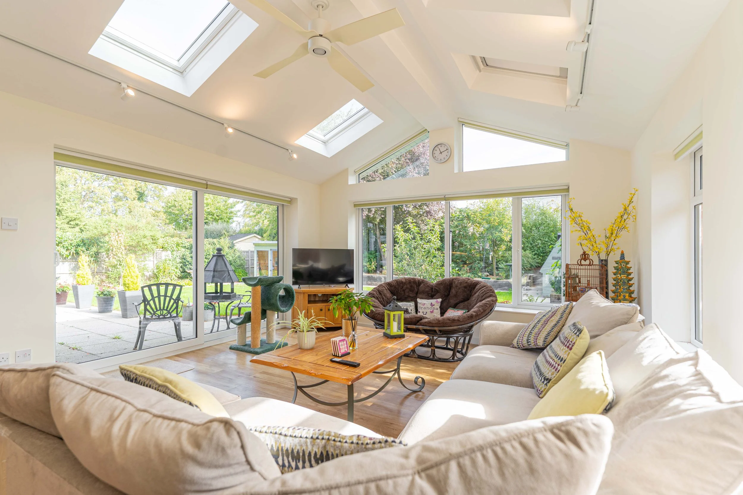 Bright living room with large glass windows and sliding door to an outdoor patio, featuring a beige sectional sofa with throw pillows, a wooden coffee table, a brown papasan chair, a cat tree, a TV, and decorative plants, with a ceiling fan and skyli