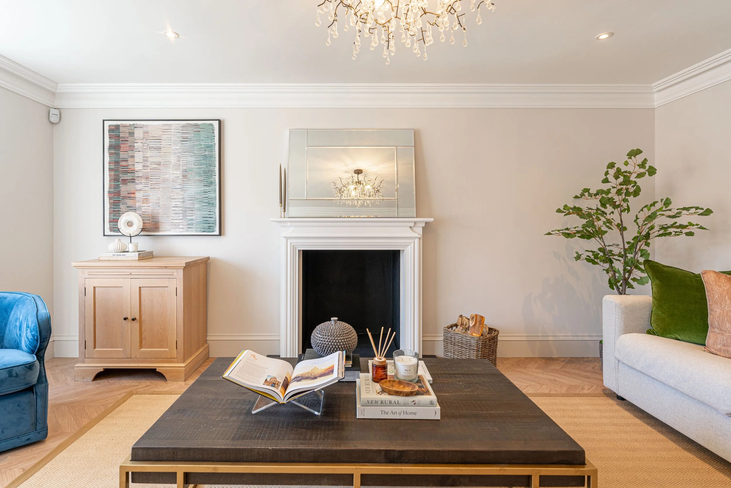 Living room with white walls, a fireplace, abstract artwork, large mirror, chandelier, potted plant, white sofa with green and pink cushions, blue armchair, wooden cabinet, and a low table with decorative items.