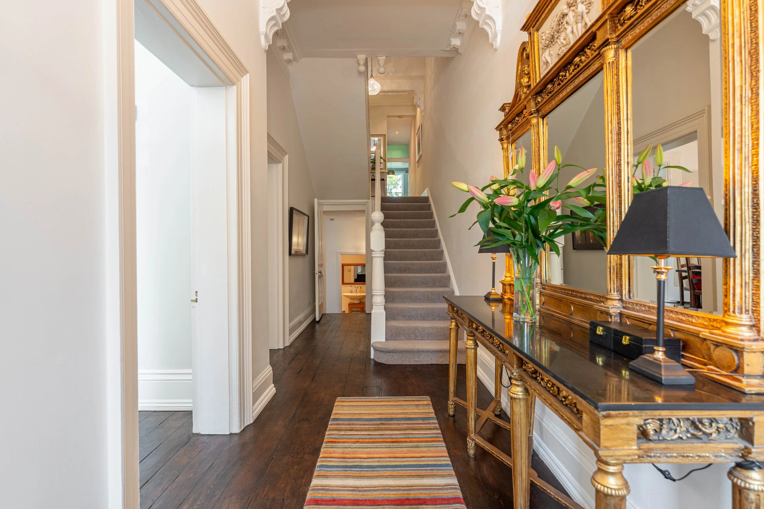 Entryway of a house with dark wood flooring, a striped rug, a gold ornate mirror, a console table with lamps and vases, a staircase with carpeted steps, and a hallway leading to other rooms.