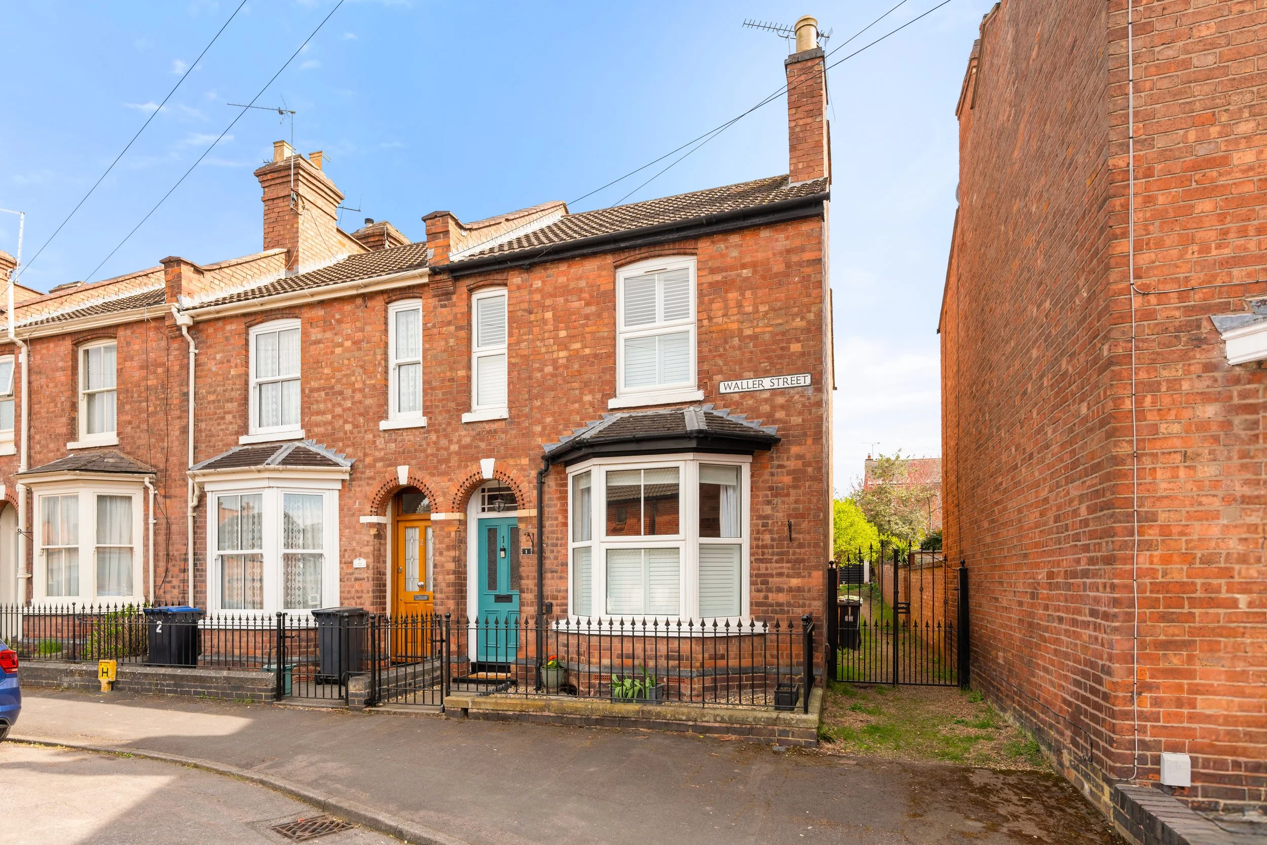 A row of brick terraced houses on Waller Street, featuring bay windows, colorful doors, small front yards with plants, and a narrow alleyway on the right side. The street is paved and has a blue sky above.