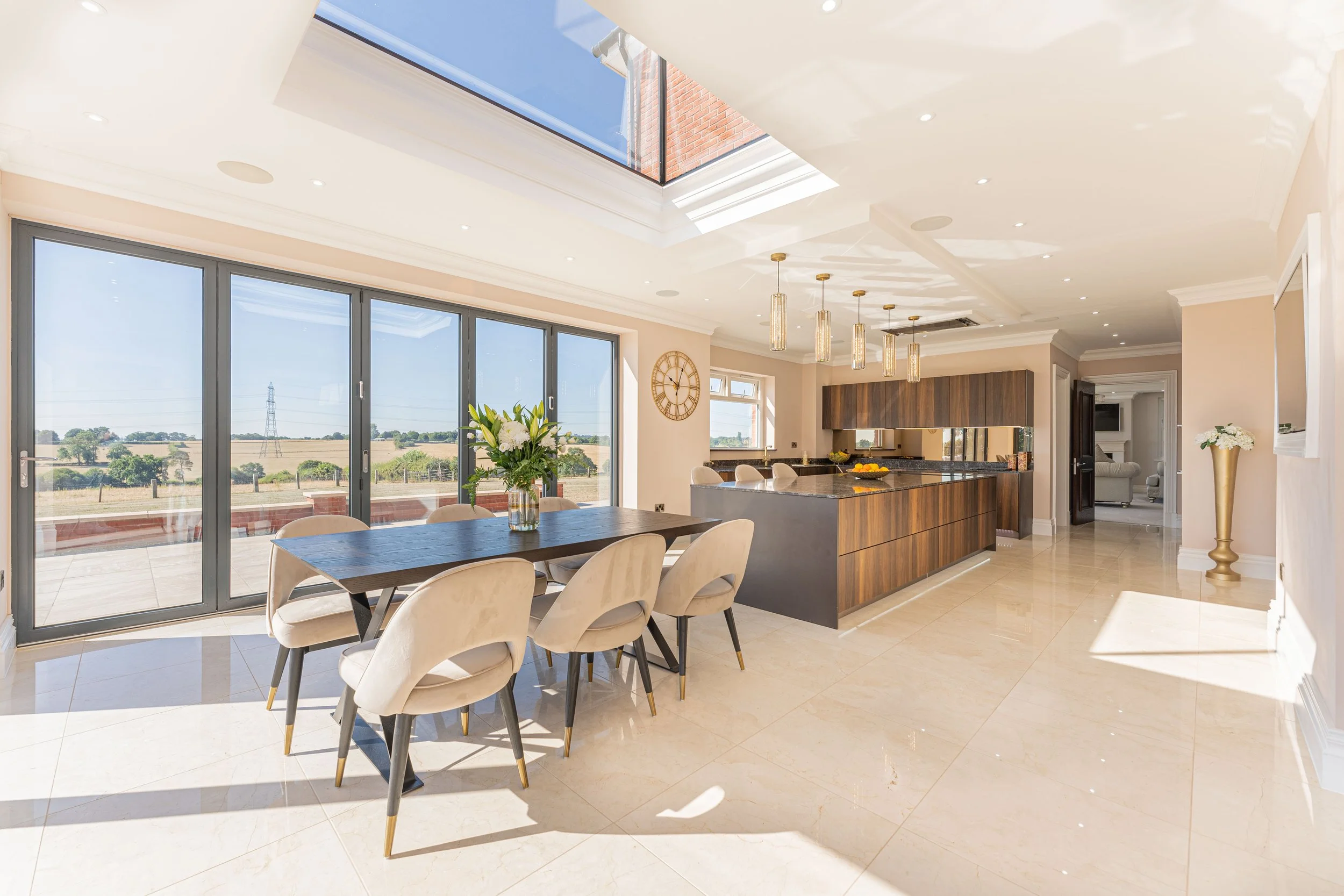 Bright open-concept kitchen and dining area with large sliding glass doors, a dark wood dining table with cream chairs, a central island with pendant lighting, and expansive skylights letting in natural light.