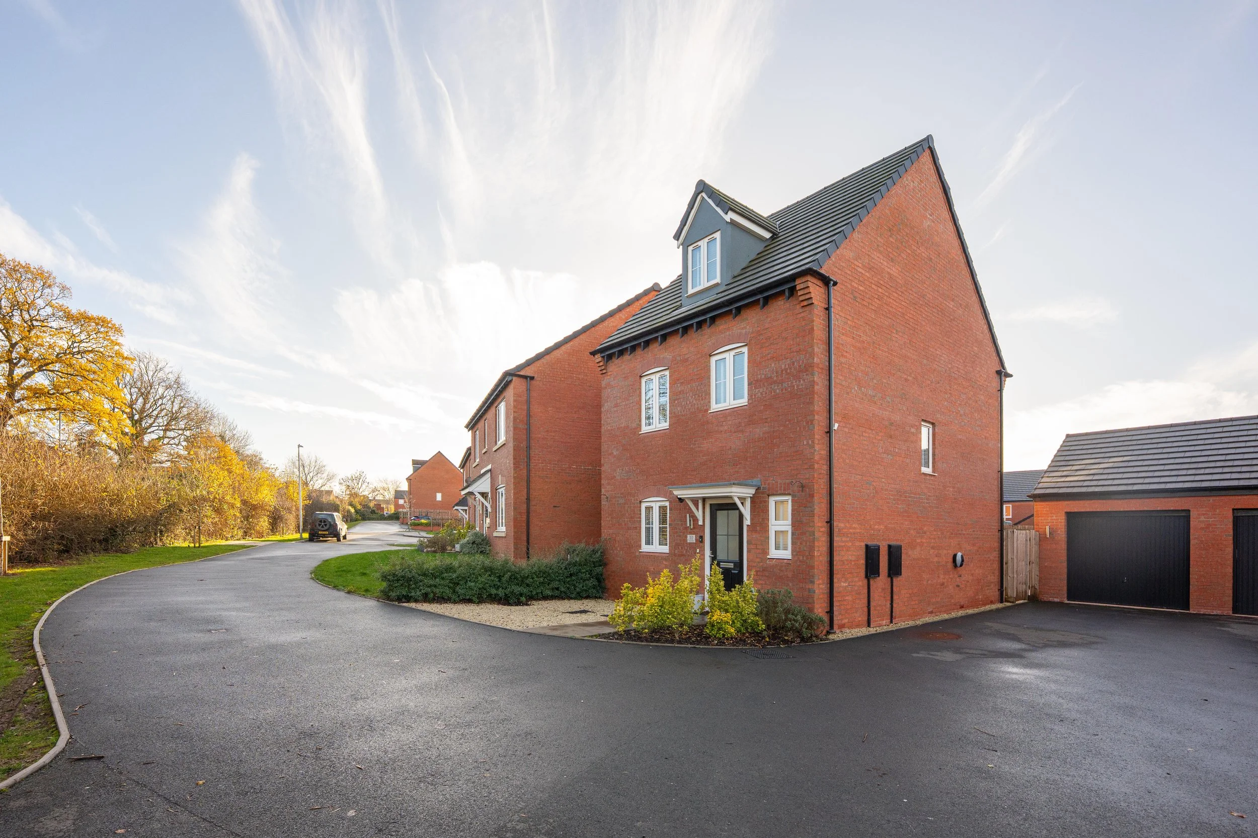 A modern brick house with a black door, white-framed windows, and a small front garden, situated on a curved asphalt street in a suburban neighborhood under a partly cloudy sky.