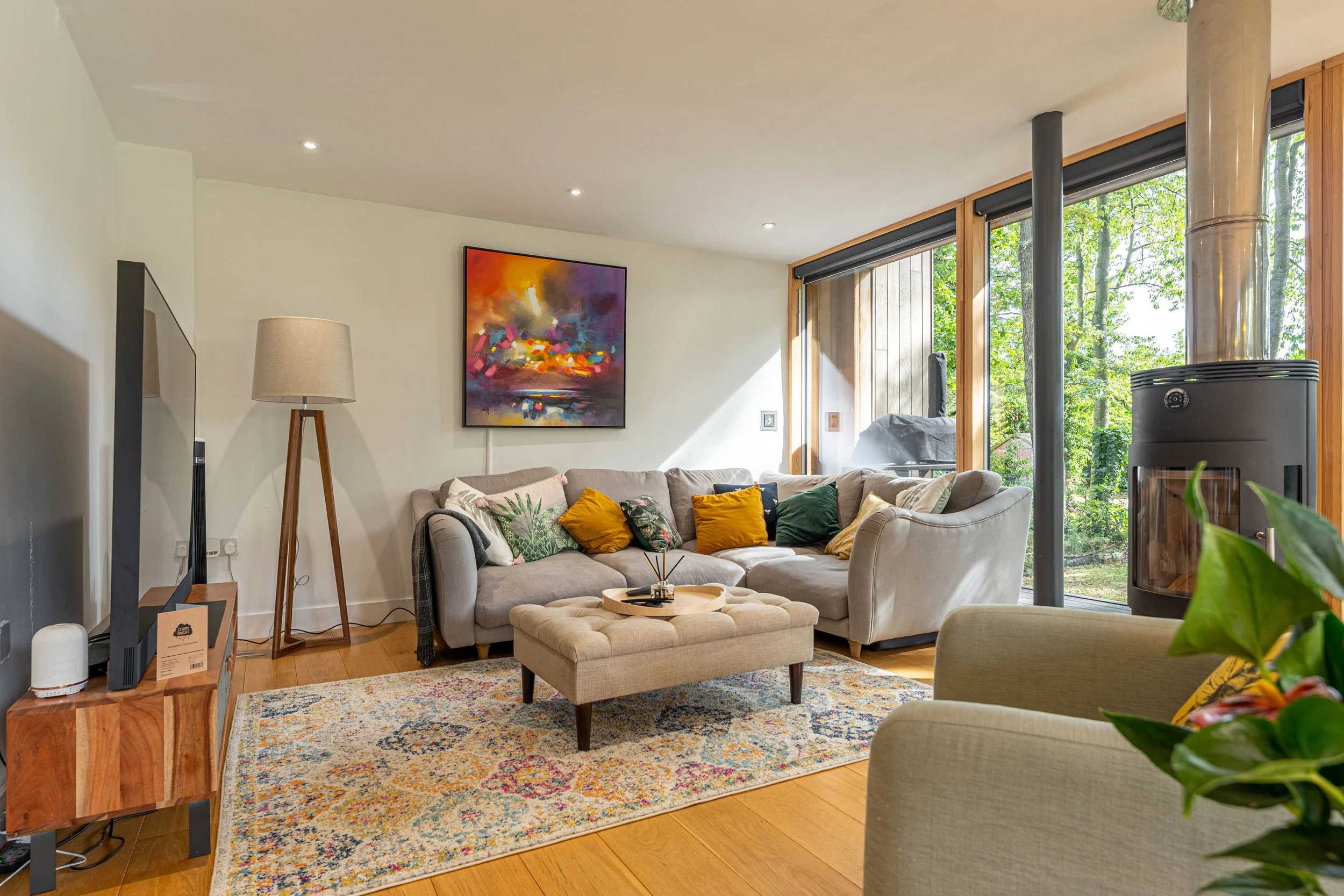 Living room with beige sectional sofa, colorful cushions, a rectangular ottoman, a patterned rug, large windows with views of greenery, a painting on the wall, a floor lamp, a wood tv stand, a fireplace, and a potted plant.