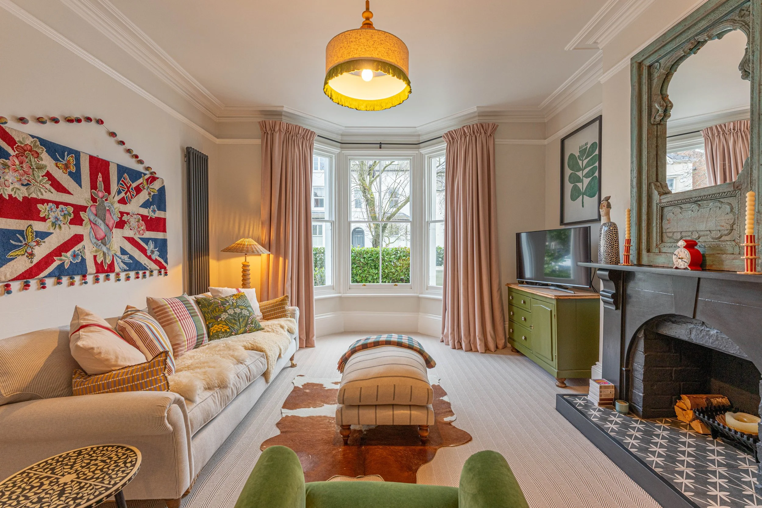 Living room with a beige sofa, colorful pillows, a cowhide rug, a large bay window with pink curtains, a black fireplace with decorative objects, a green cabinet with a TV, and a ceiling light.
