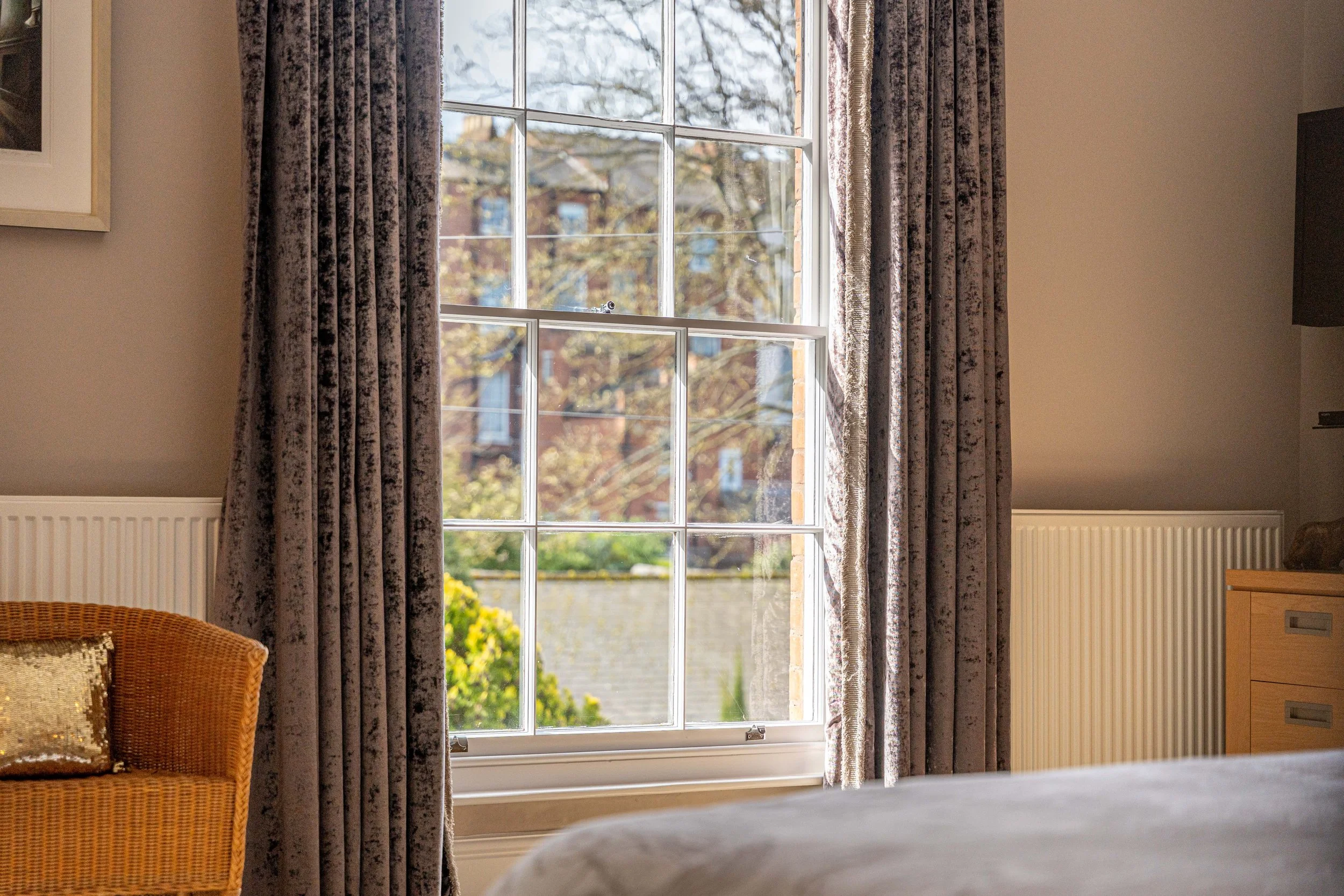 A view through a large window with grid panels, showing trees, houses, and a blue sky outside. Inside, there are beige curtains, a wicker chair with a gold pillow, a wooden dresser, and a beige wall with a picture frame.