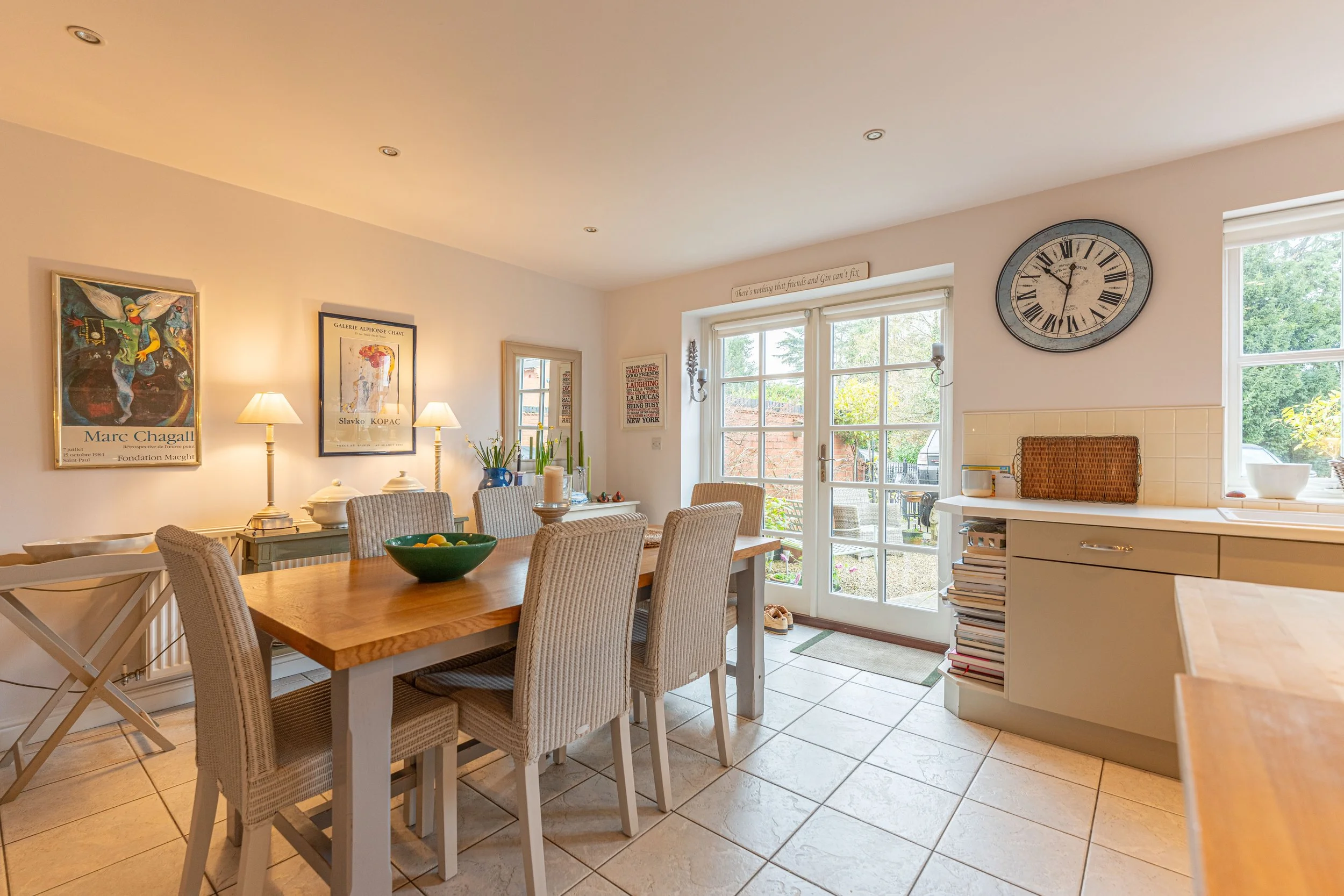 Bright dining room with a wooden table, six chairs, art posters, sideboard with lamps, large clock, and glass doors leading outside to a garden.