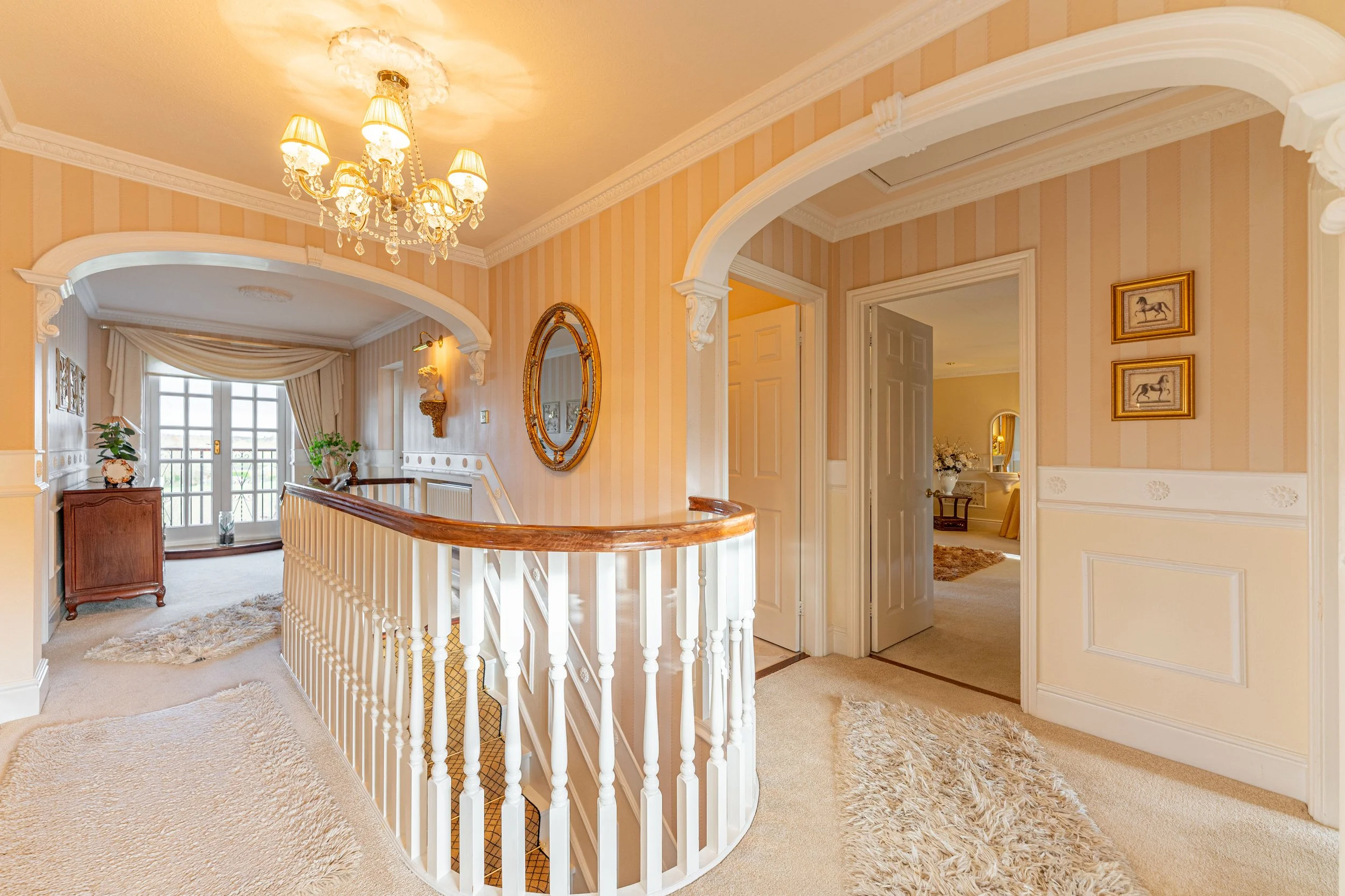 Interior of a house hallway with cream-colored walls, ornate crown molding, and a chandelier. There is a staircase with a polished wooden handrail, a decorative mirror on the wall, and a separate room visible through an open door. The area is decorat