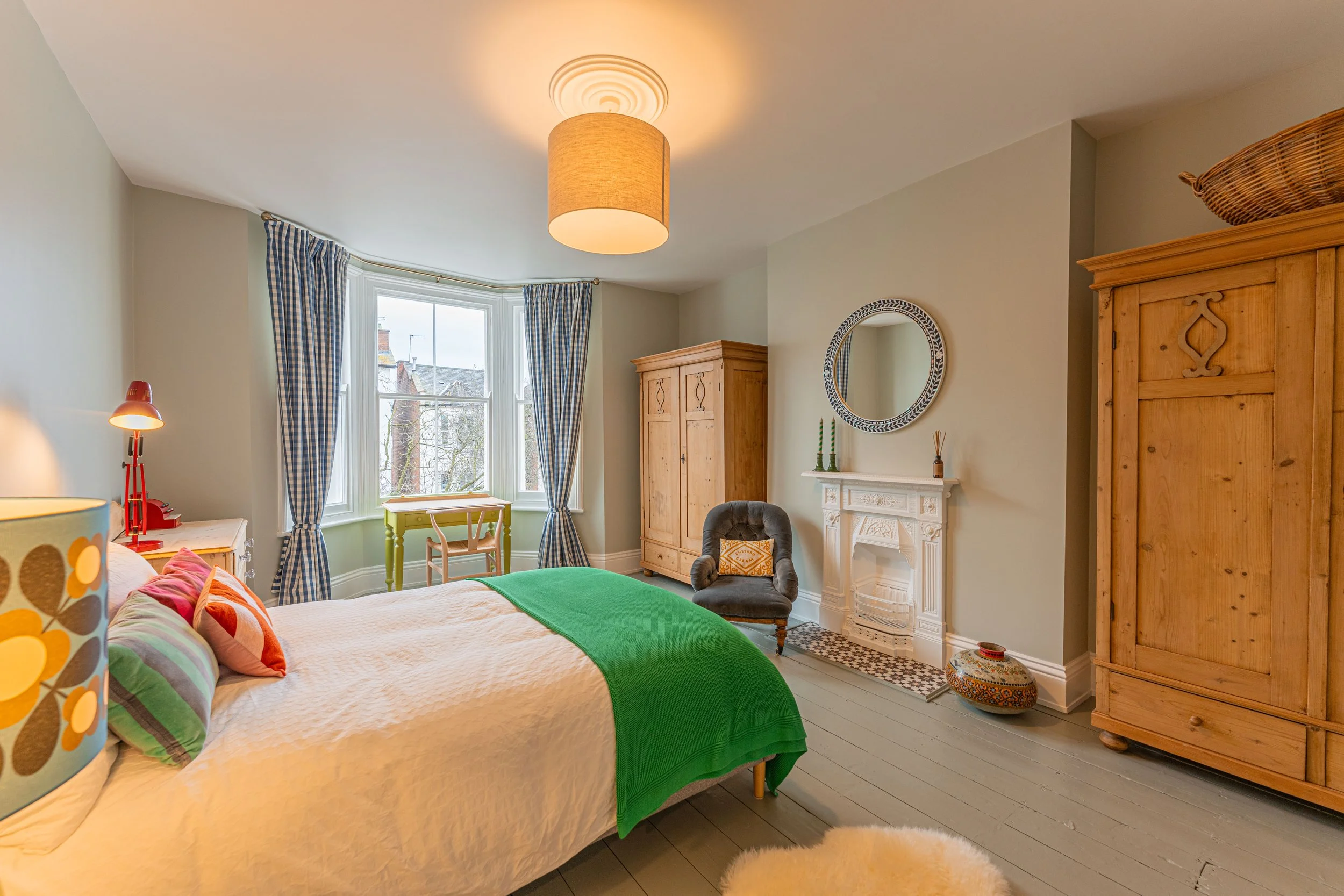 A cozy bedroom with a bed, colorful pillows, and a green blanket. There are two wooden wardrobes, a fireplace with decorative items, a gray armchair, and a small green desk by the window with blue checkered curtains. The room has a ceiling light and 