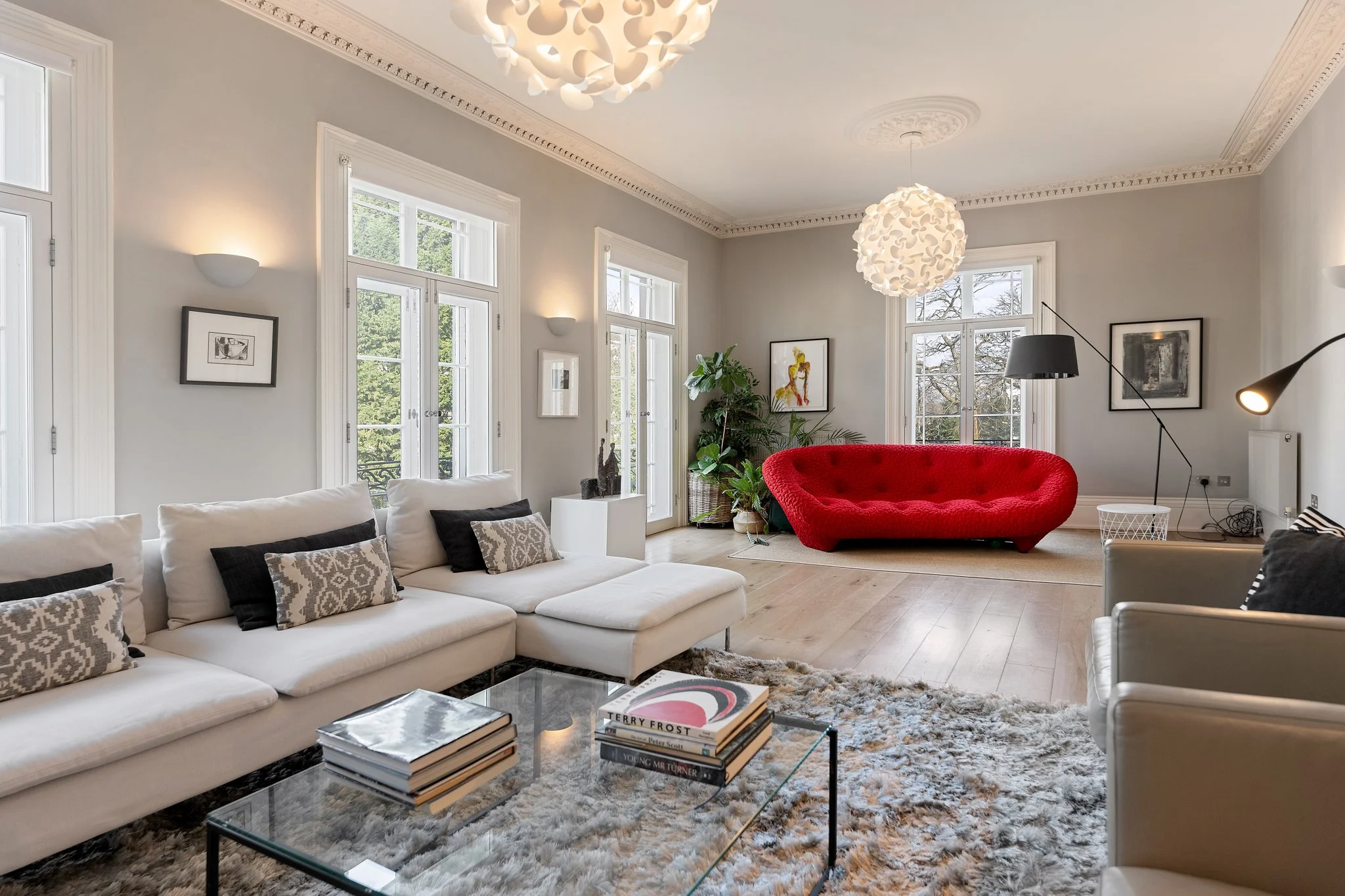 Modern living room with white sofa, glass coffee table, red velvet sofa, black and white artwork, large windows, and a textured area rug.