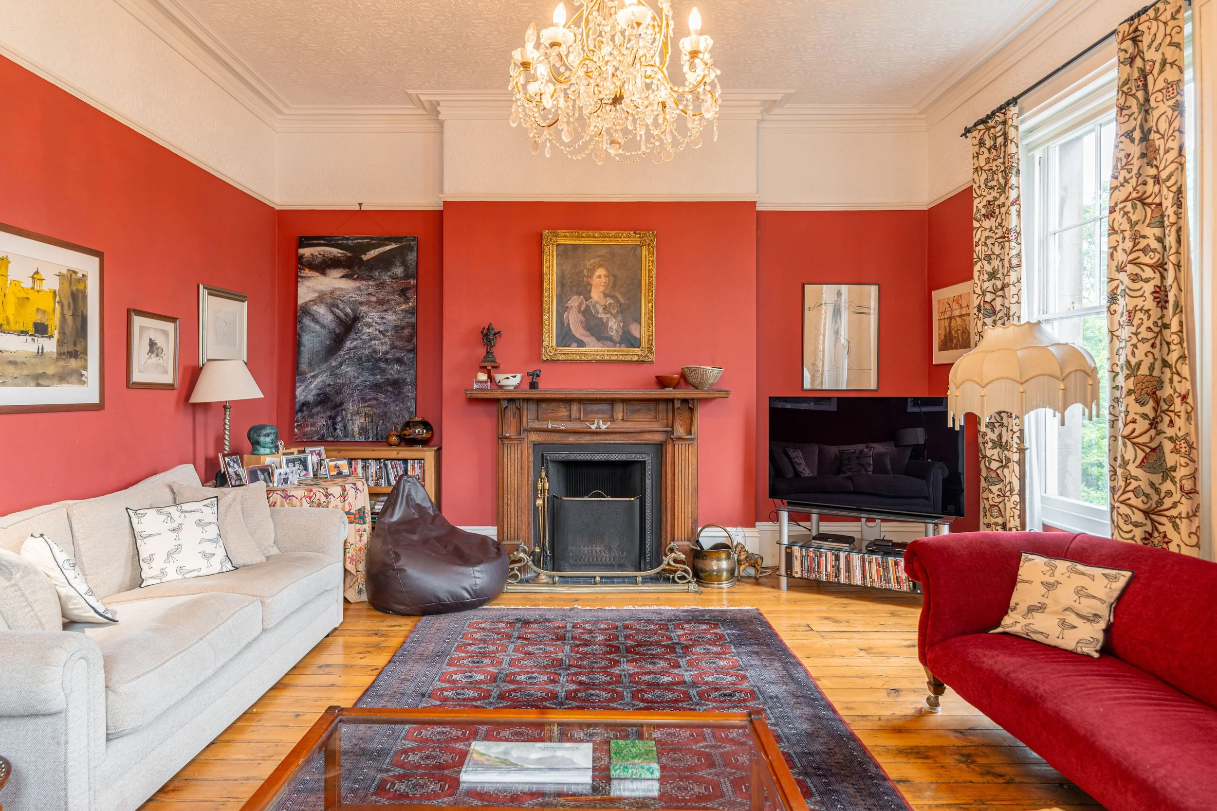 Living room with red walls, wooden floors, and a fireplace. There are two sofas—one beige and one red—with pillows, a large chandelier overhead, curtains on large windows, and various artworks on the walls. A coffee table is in the foreground, and a 