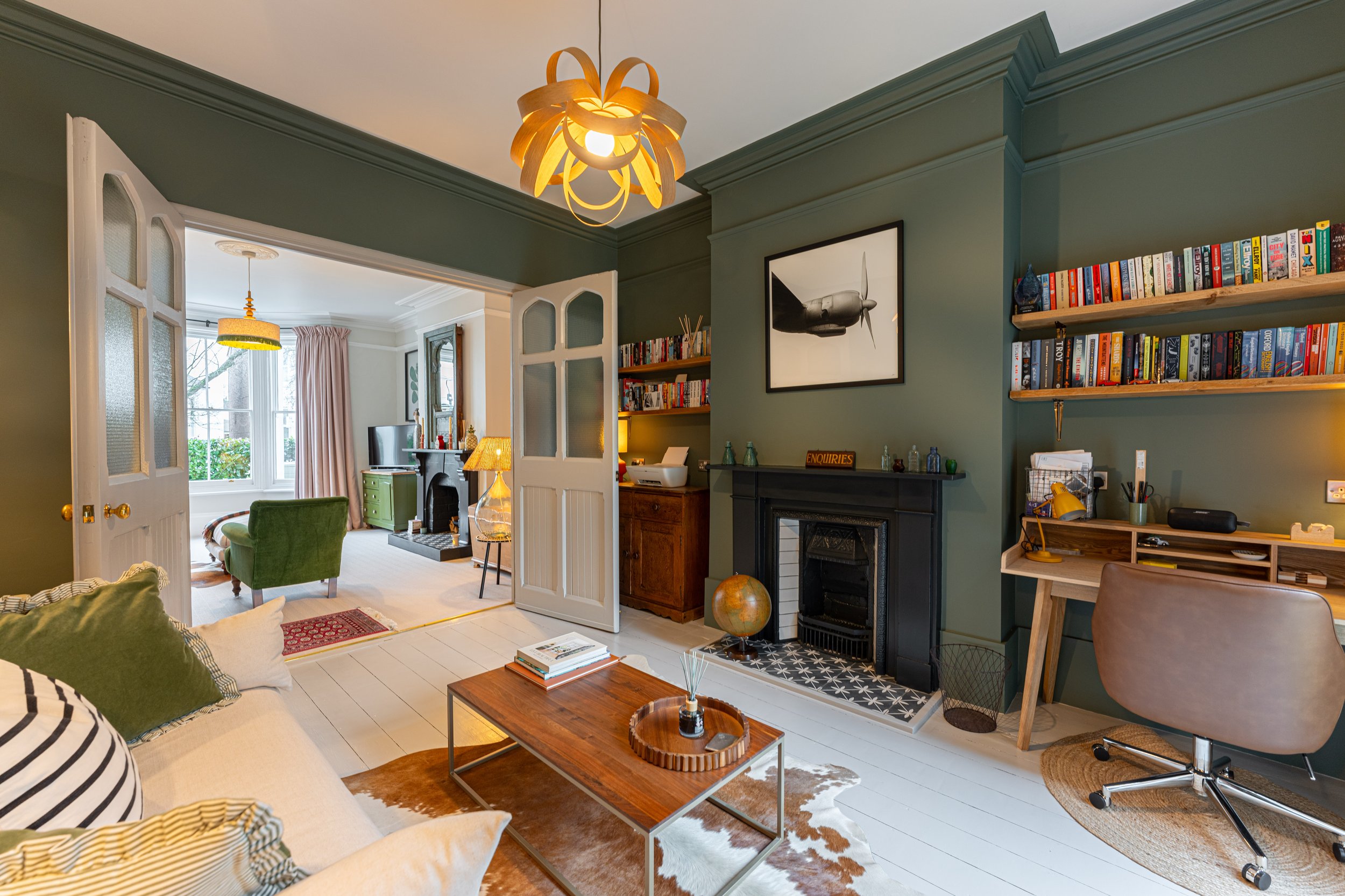 Living room with green walls, black fireplace, bookshelf, desk, and a brown armchair, view into a bright sitting area with window and pink curtains.