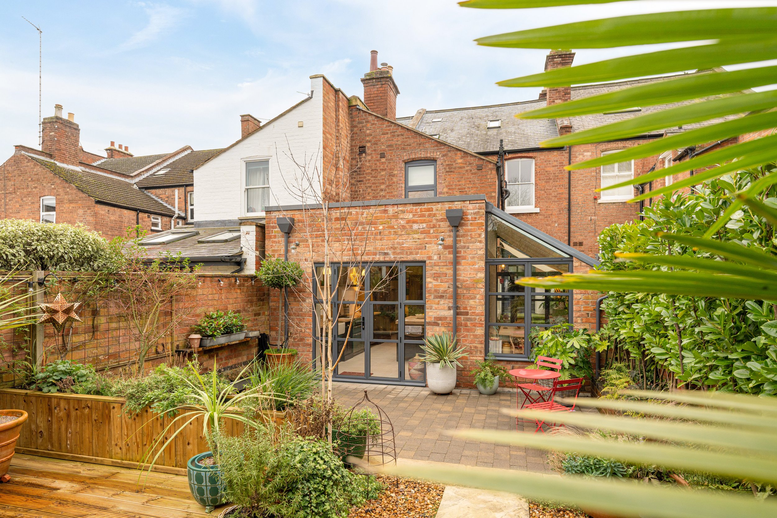 Backyard patio of a brick house with large windows and potted plants, surrounded by a fence and lush greenery, under a partly cloudy sky.