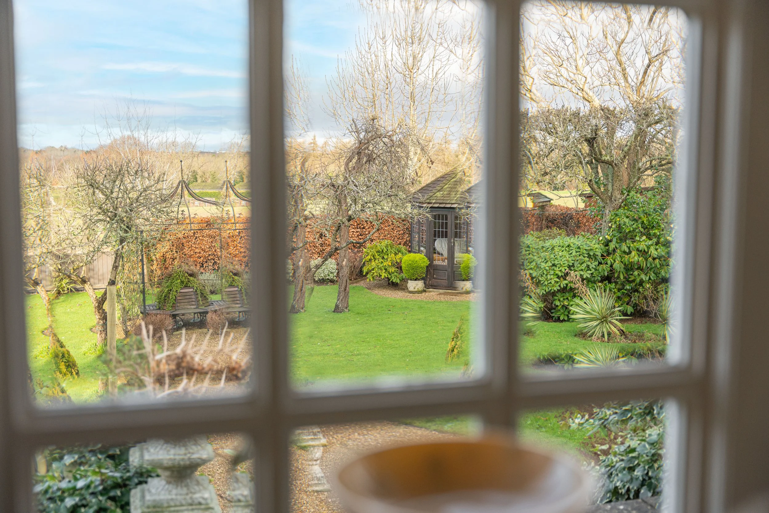View of a garden through a window with four panes, showing trees, bushes, a shed, a bench, and a well-maintained lawn.