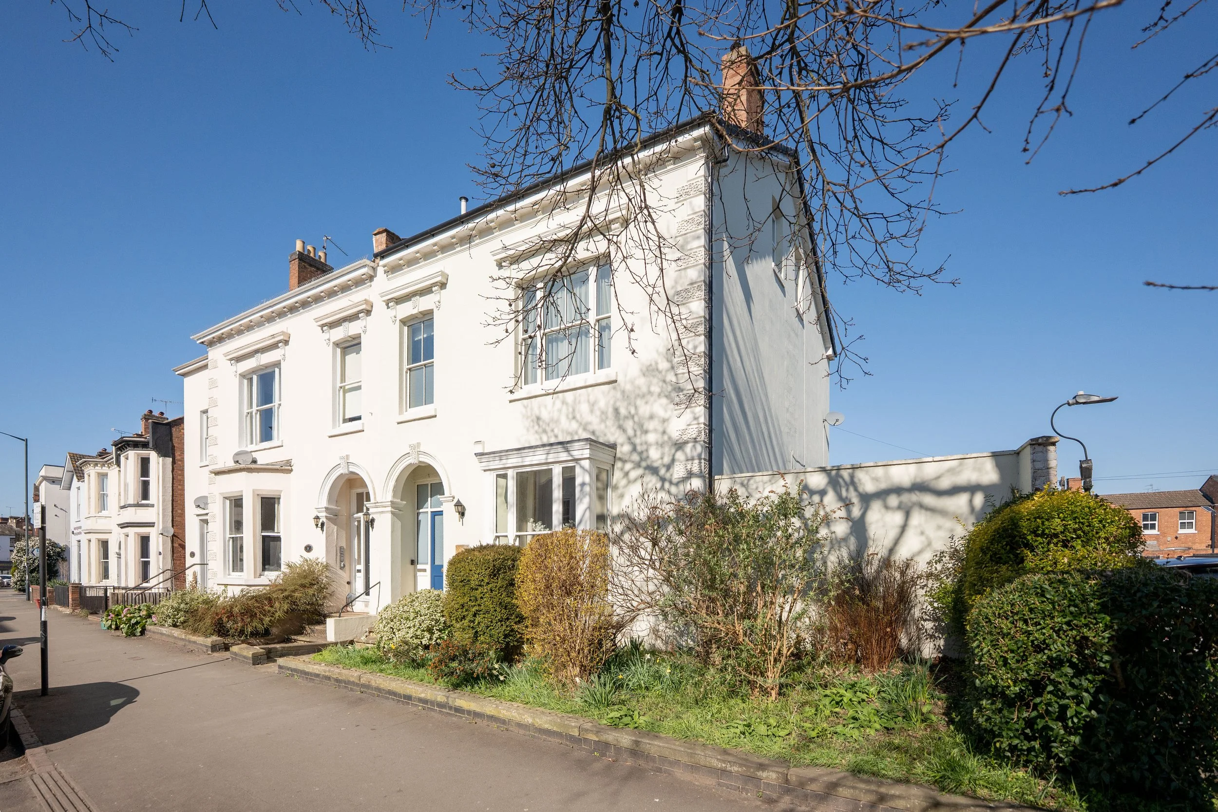 A white row house with bay windows on a quiet street in sunlight, with some bushes and trees in front, and a clear blue sky overhead.