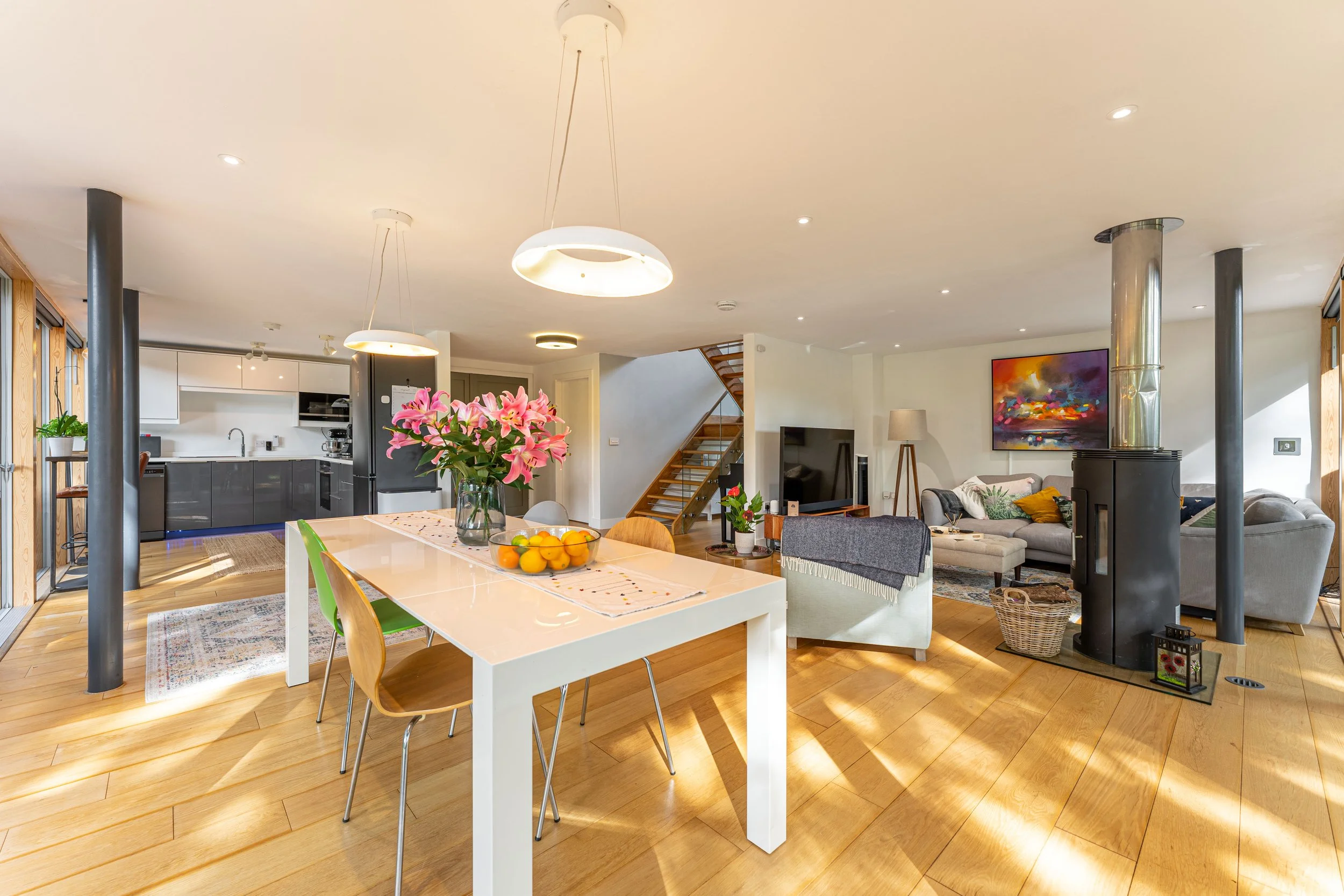 Open-concept living and dining area with wooden flooring, a white dining table with pink flowers in a vase, gray and beige sofas, a wall-mounted TV, a staircase, and kitchen in the background with white cabinets and black appliances.