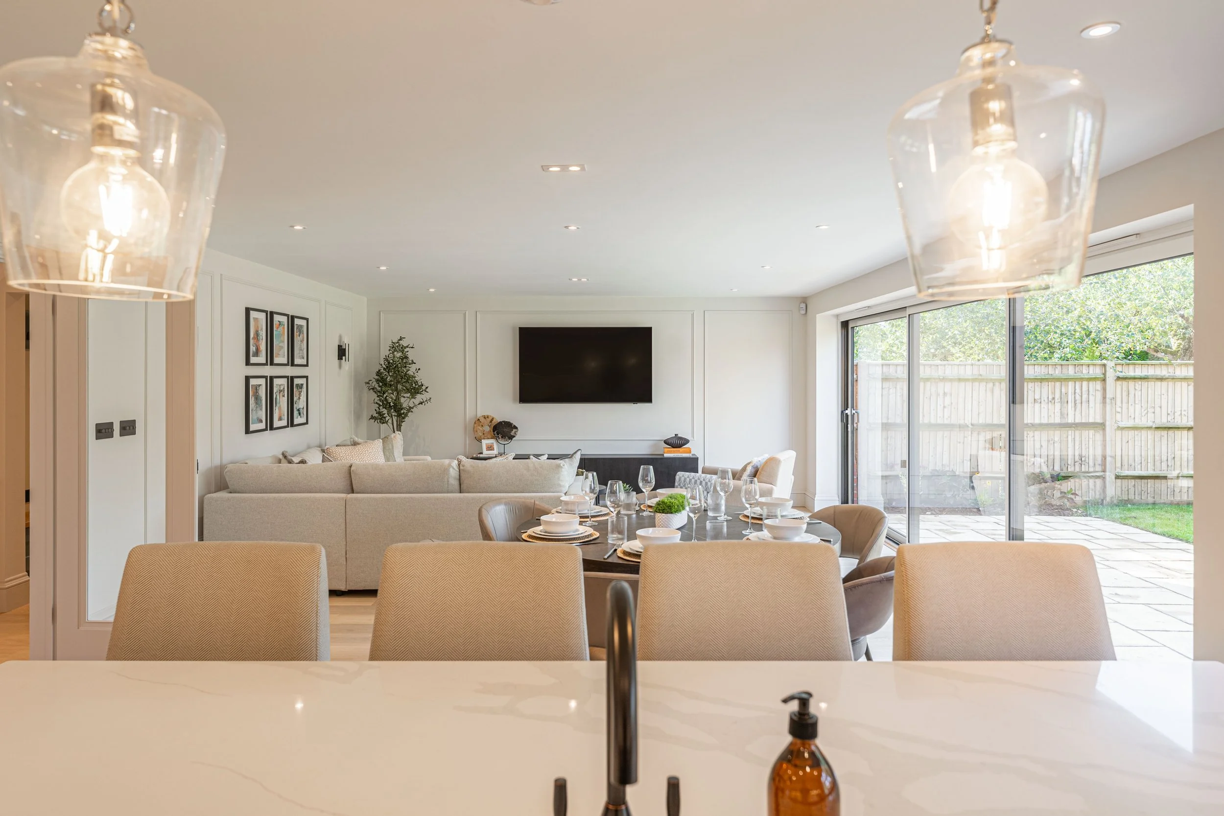 Modern open-concept living and dining area with large sliding glass doors to the backyard, a mounted flat-screen TV, a round dining table set with plates and glasses, beige chairs, hanging pendant lights, and a white kitchen counter with a soap dispe