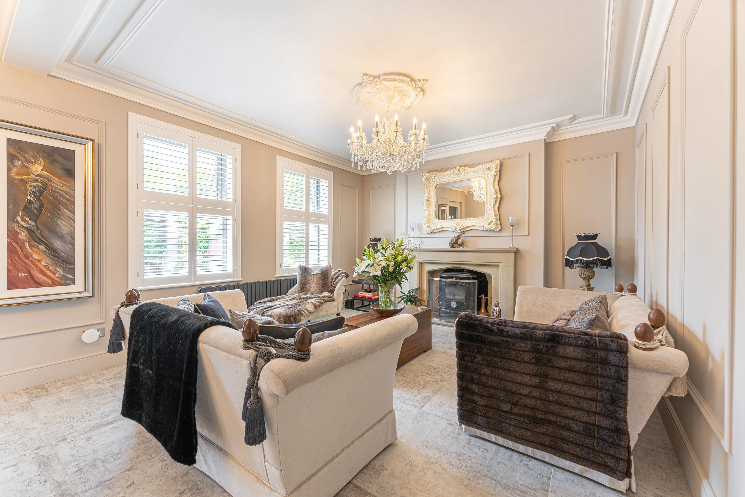 Elegant living room with cream and brown sofas, a fireplace with a mirror above, a chandelier, and a vase of white flowers on a coffee table.