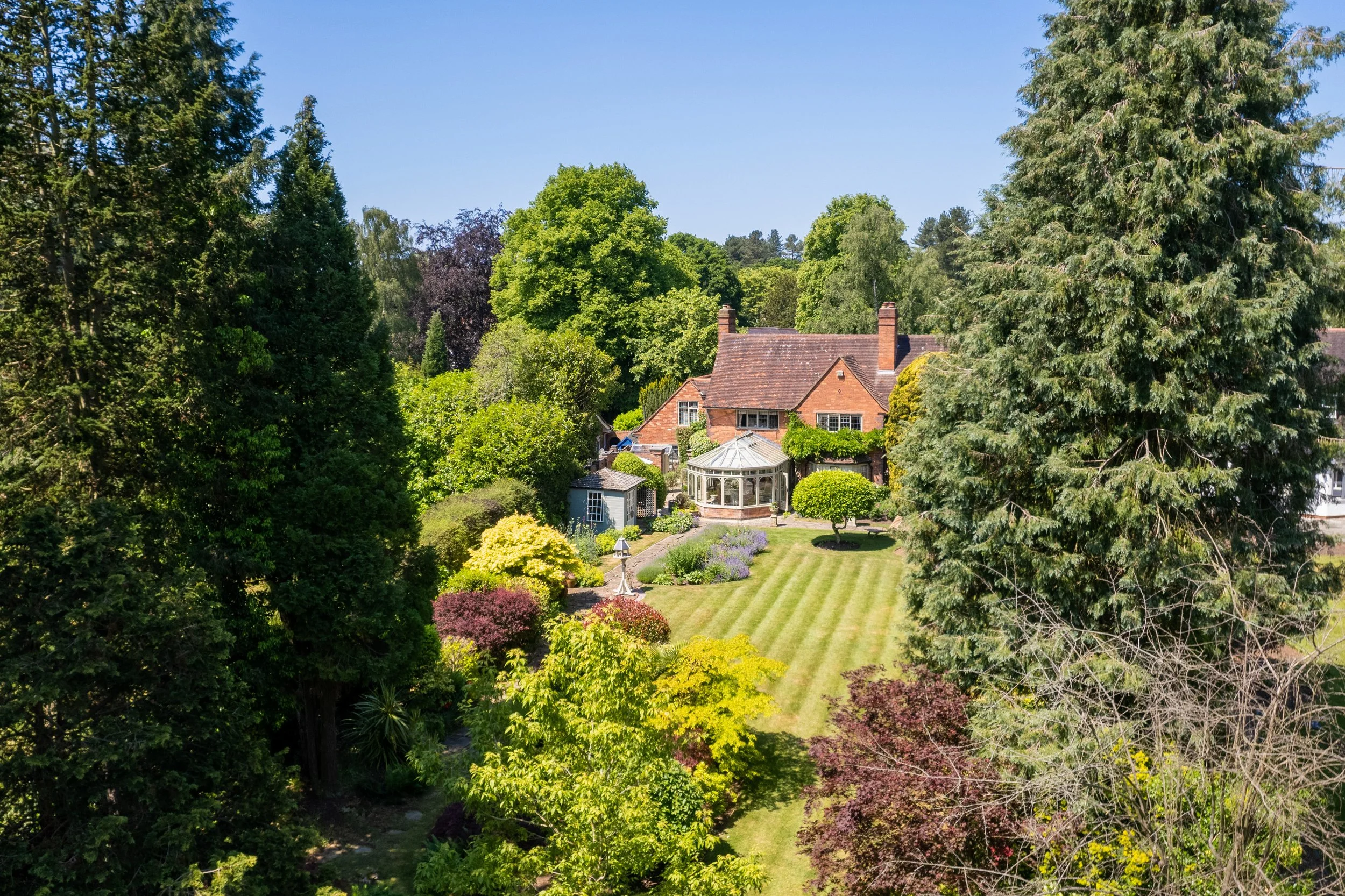 A large brick house surrounded by lush green trees and a well-manicured lawn with striped grass and various colorful shrubs.