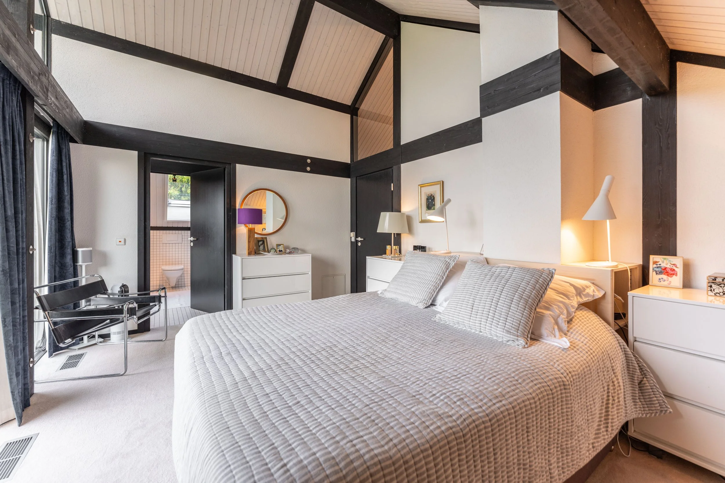 A bedroom with a large bed, white furniture, and a visible bathroom door. The room has black and white walls with a sloped ceiling, a sitting chair, and personal items on nightstands and dressers.