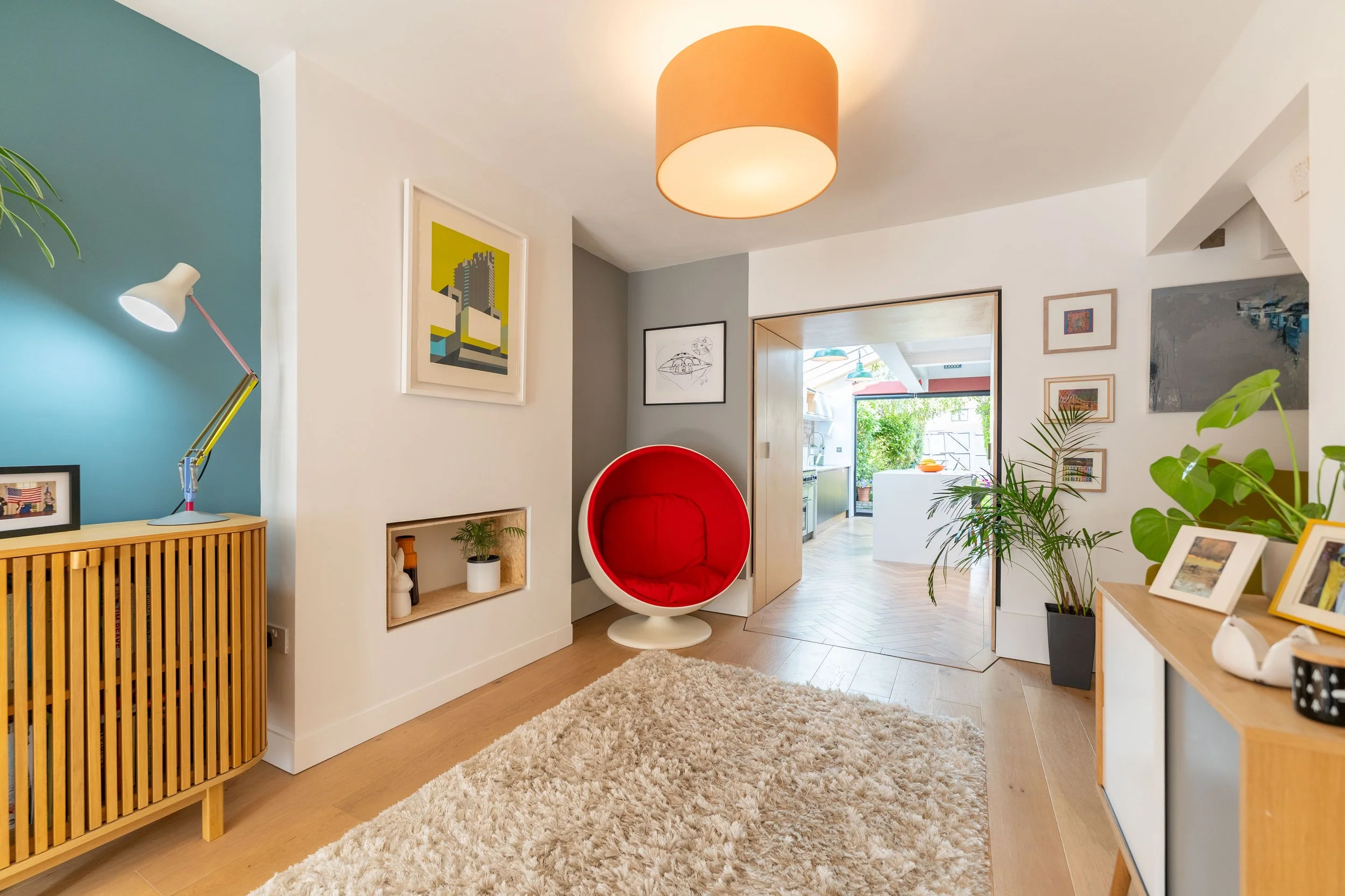 Bright living room with wooden floors, a beige fluffy rug, a red modern bubble chair, and various framed artwork on white and gray walls. There's a side cabinet with a table lamp, and a large potted plant. The room opens into a bright kitchen area.