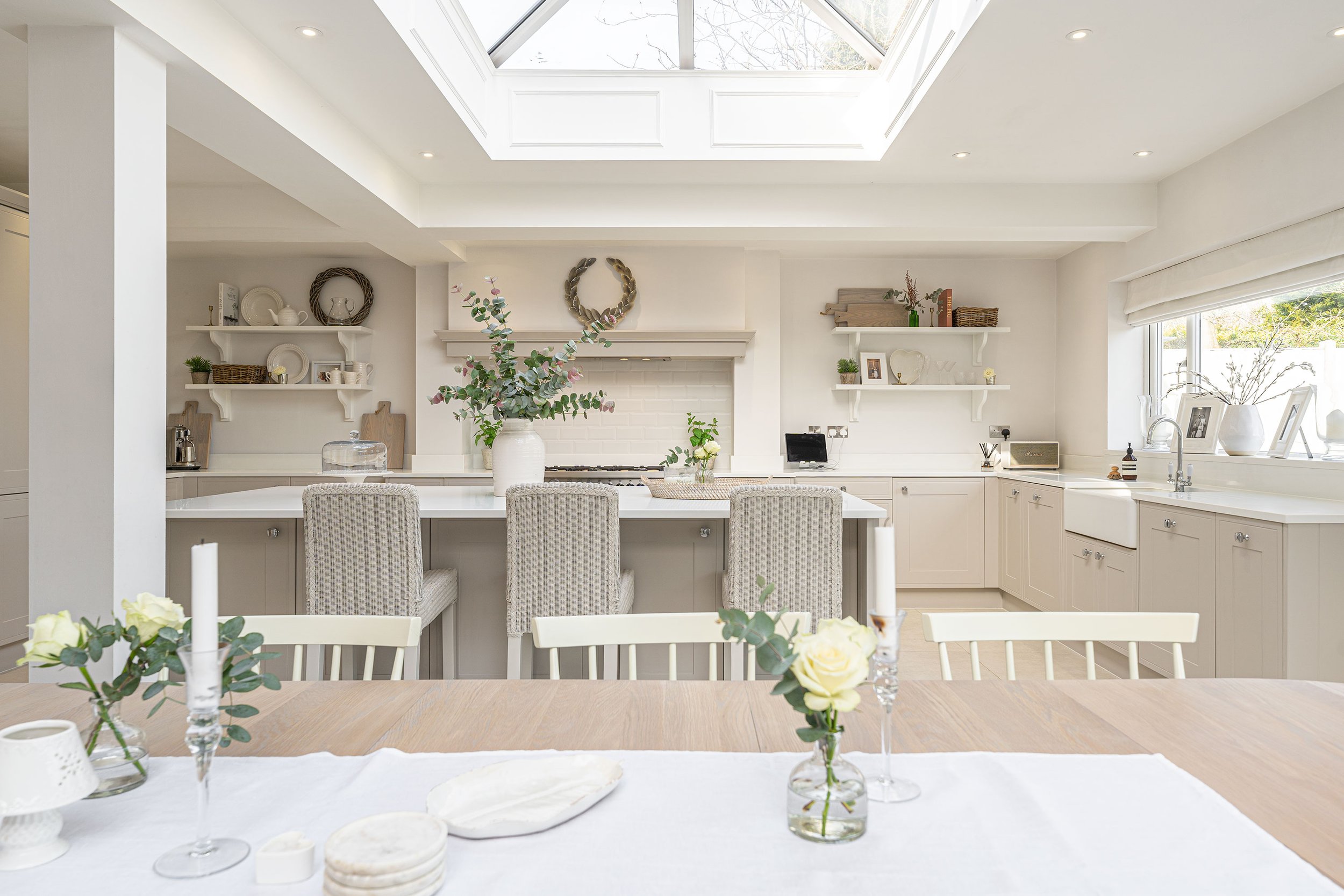 Bright kitchen with skylight, white cabinets, open shelving, dining table with white chairs, and decorative vases with flowers.