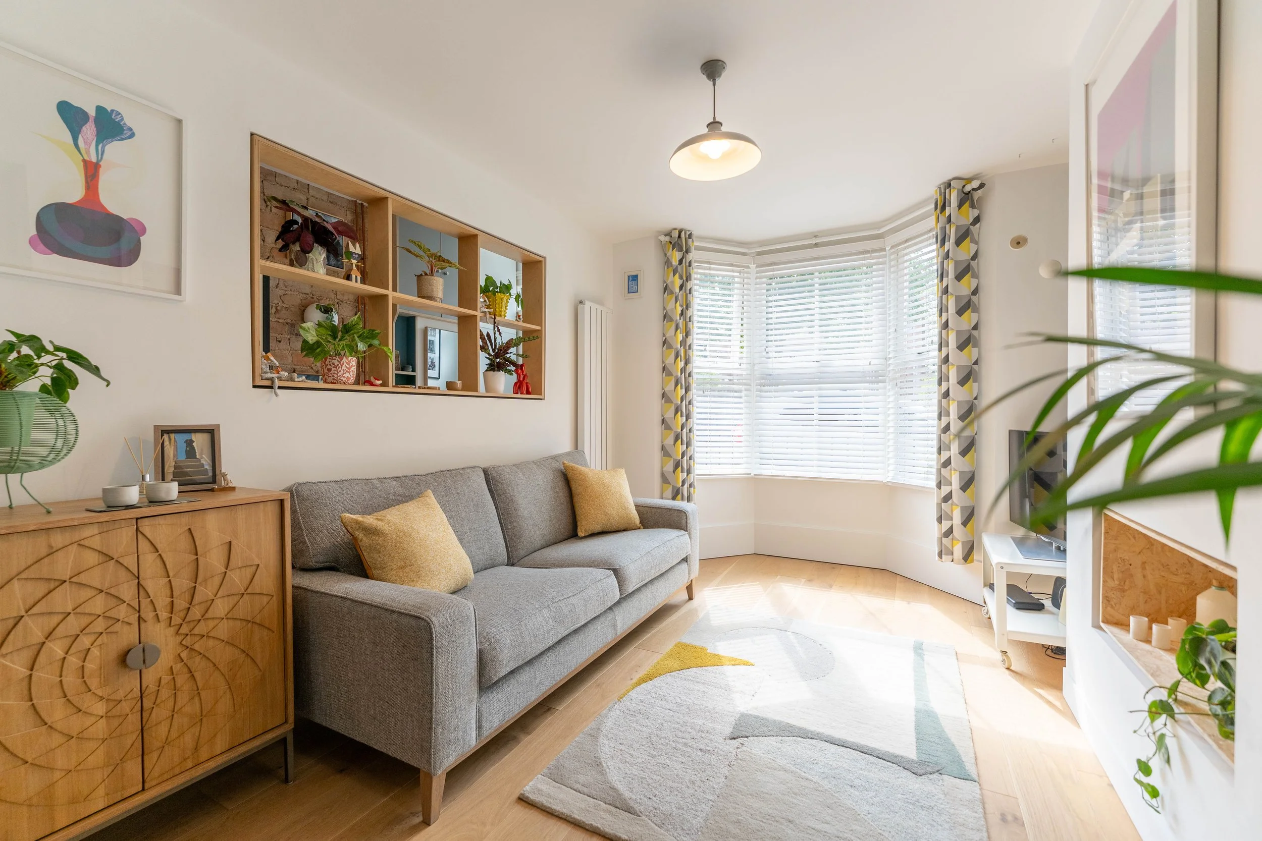 Bright living room with a gray sofa with two beige pillows, wooden sideboard with plants, a wall shelf with decorative items, large bay window with patterned curtains, colorful modern rug, and a television in the corner.