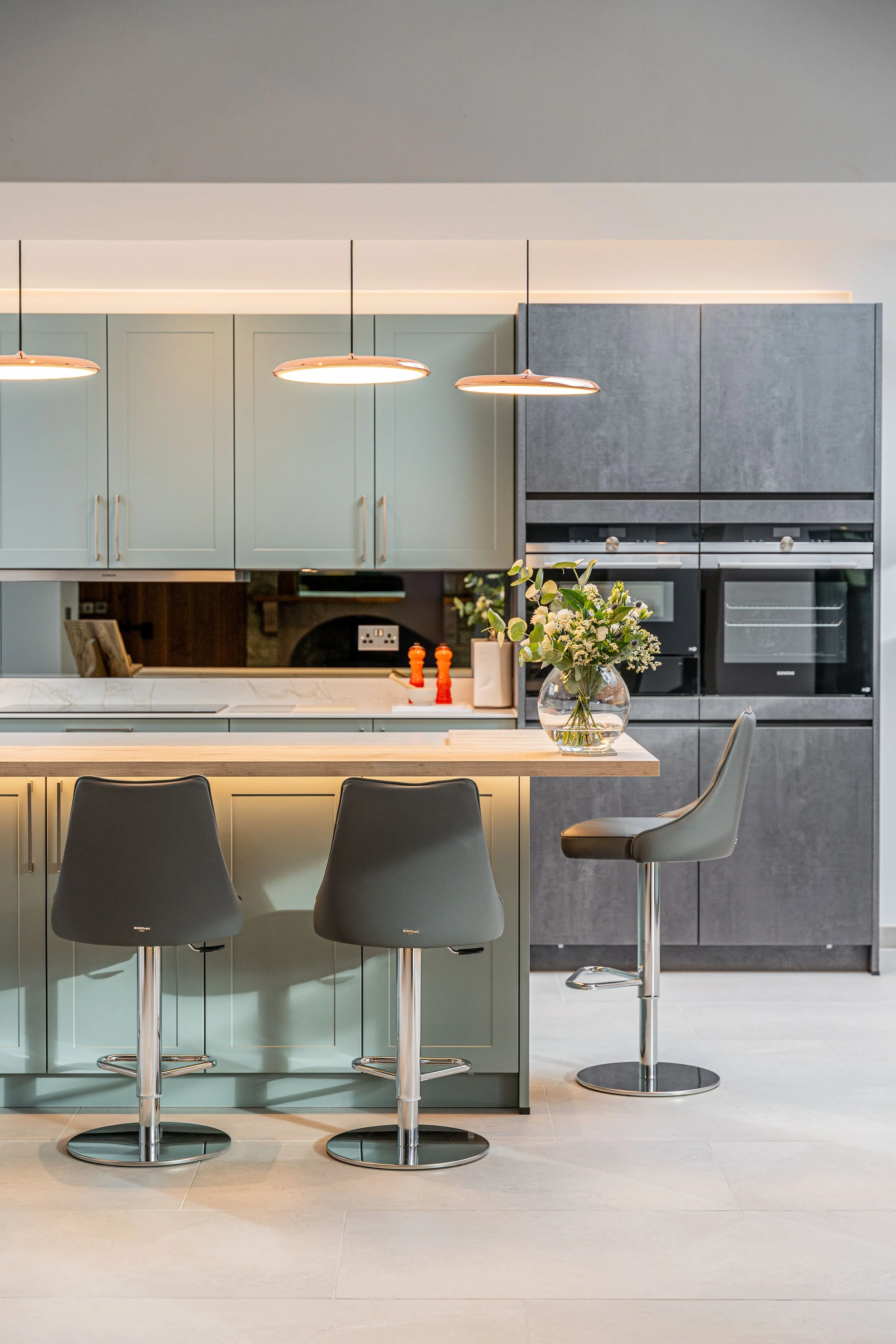 Modern kitchen with pastel green and dark gray cabinets, pendant lights, a kitchen island with bar stools, and a vase of flowers on the counter.