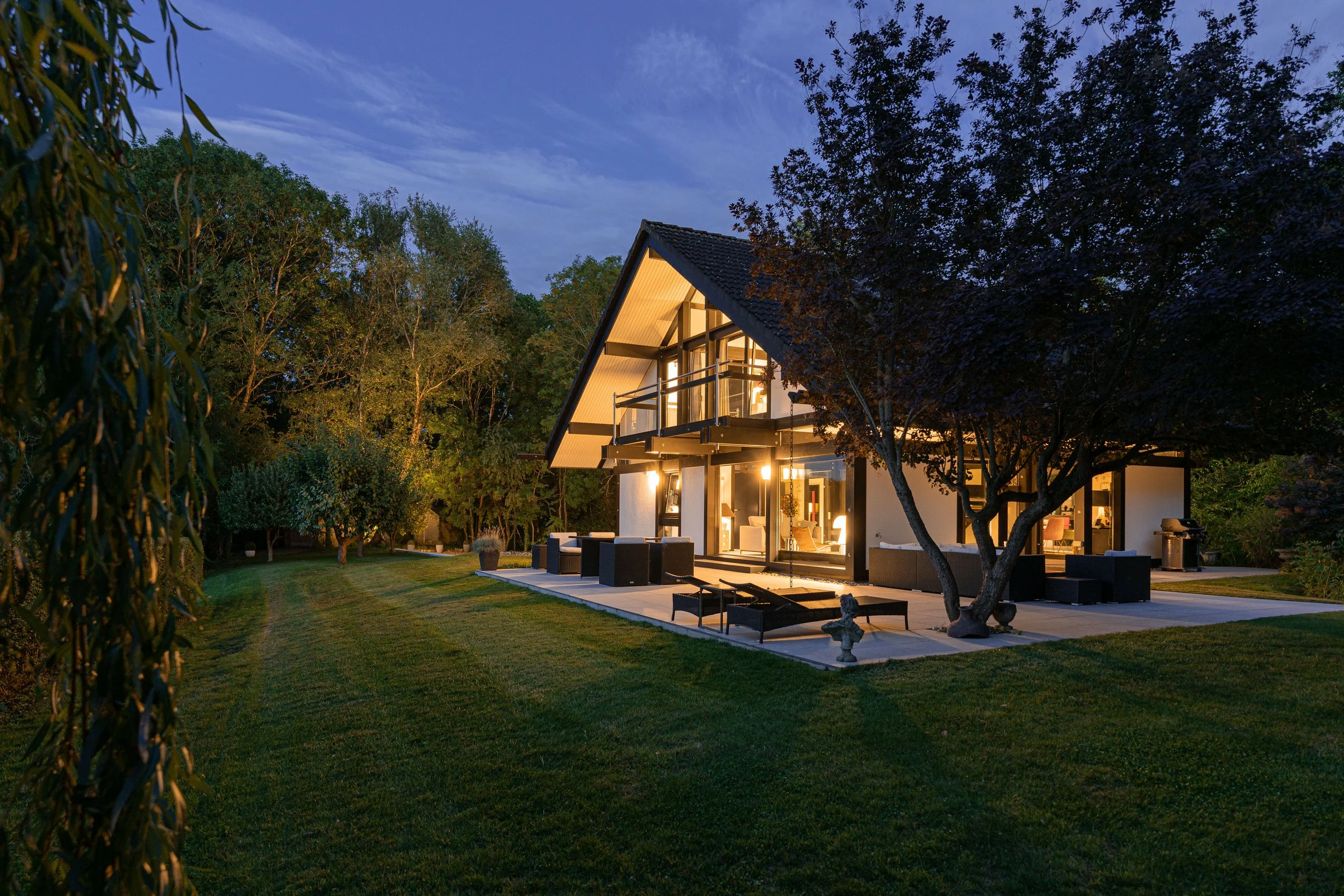 A modern house with large glass windows and a pitched roof, illuminated inside, surrounded by a well-manicured lawn and trees at dusk.