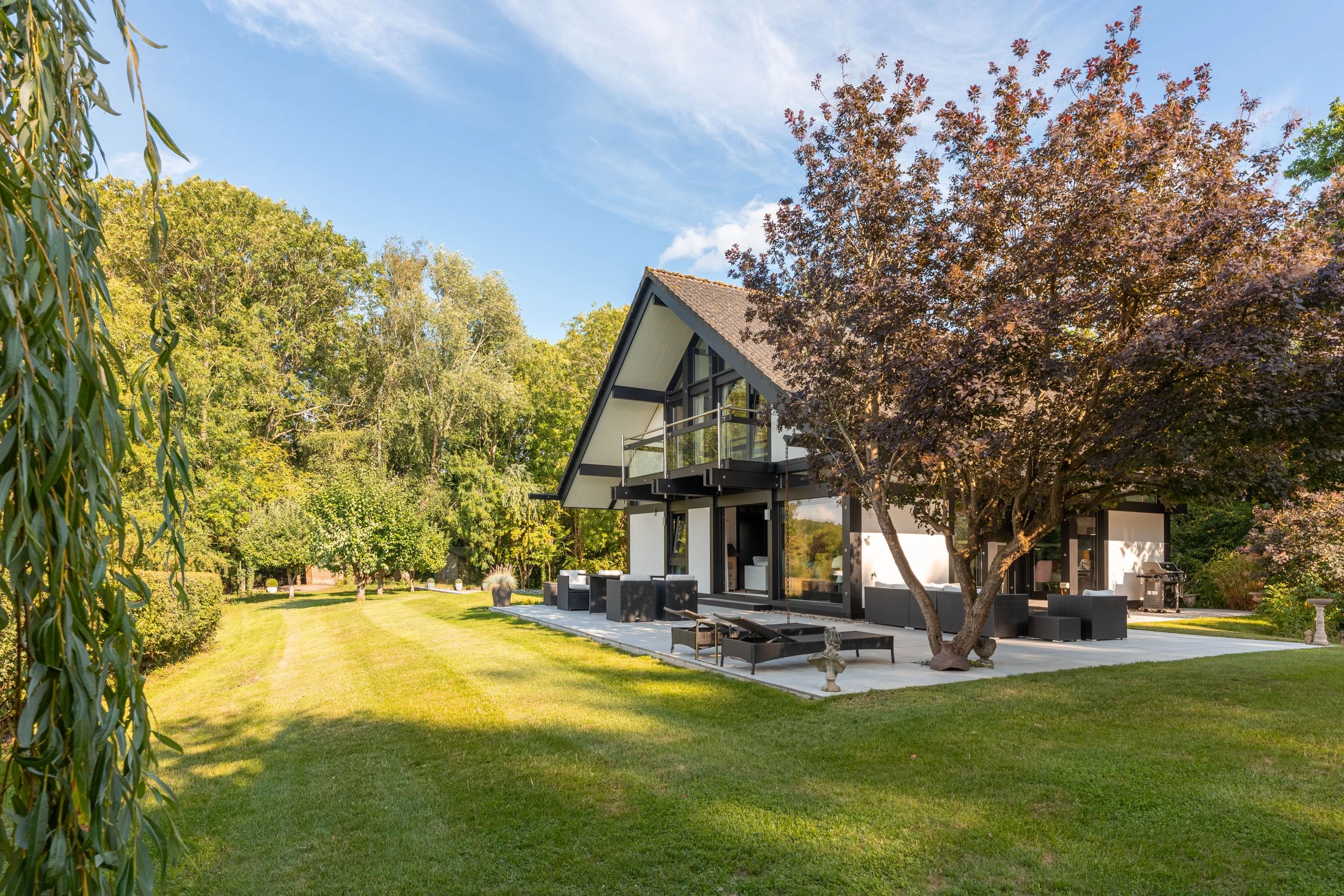 Modern house with large backyard, patio, outdoor seating, trees, and lush green grass under a blue sky.