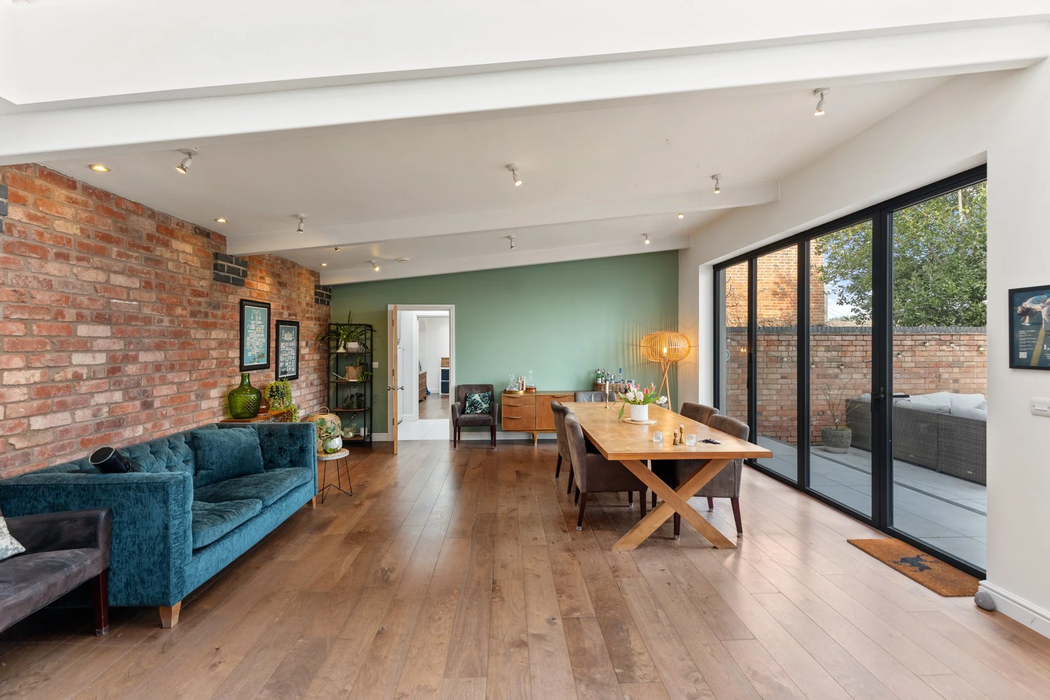 Open plan living and dining area with hardwood floors, a brick wall, a large window with a sliding door leading to an outdoor patio, a blue velvet sofa, a black quilted sofa, a wooden dining table with chairs, a standing lamp, and a green accent wall