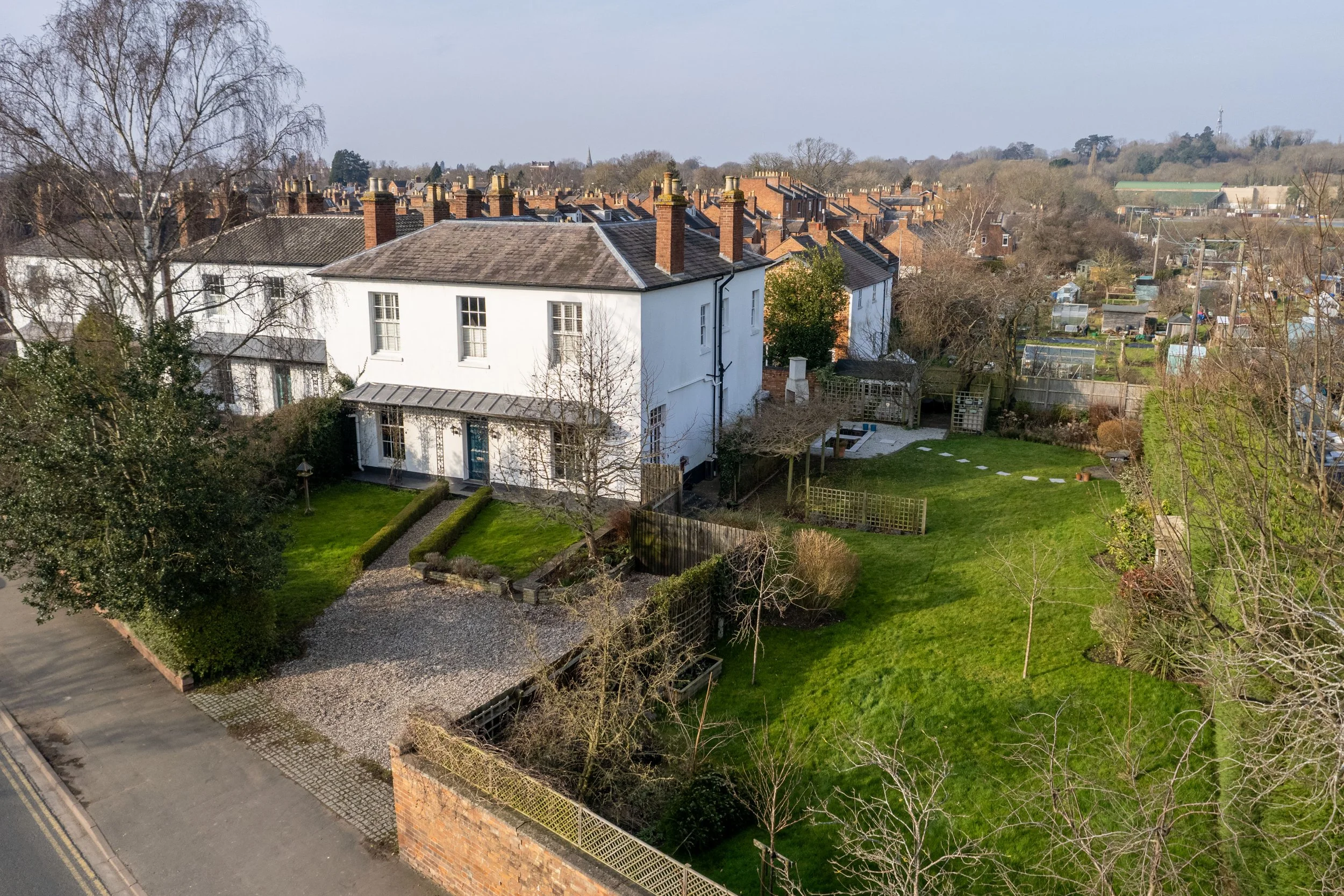 Aerial view of a white multi-story house with a garden, trees, and a gravel driveway, in a neighborhood with rooftops and greenhouses in the background during daytime.