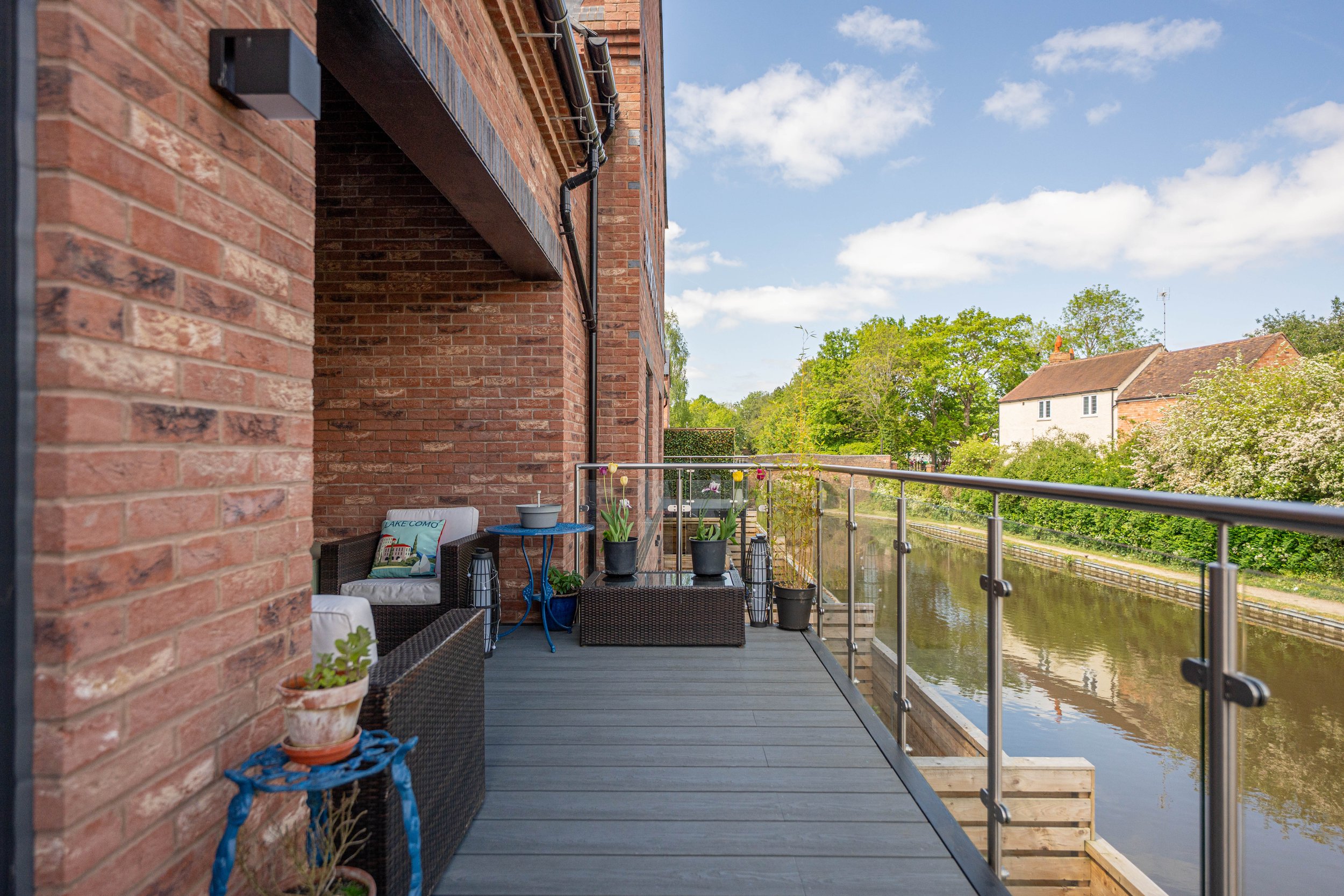 Balcony with outdoor furniture and potted plants overlooking a waterway, with trees and houses in the background on a sunny day.