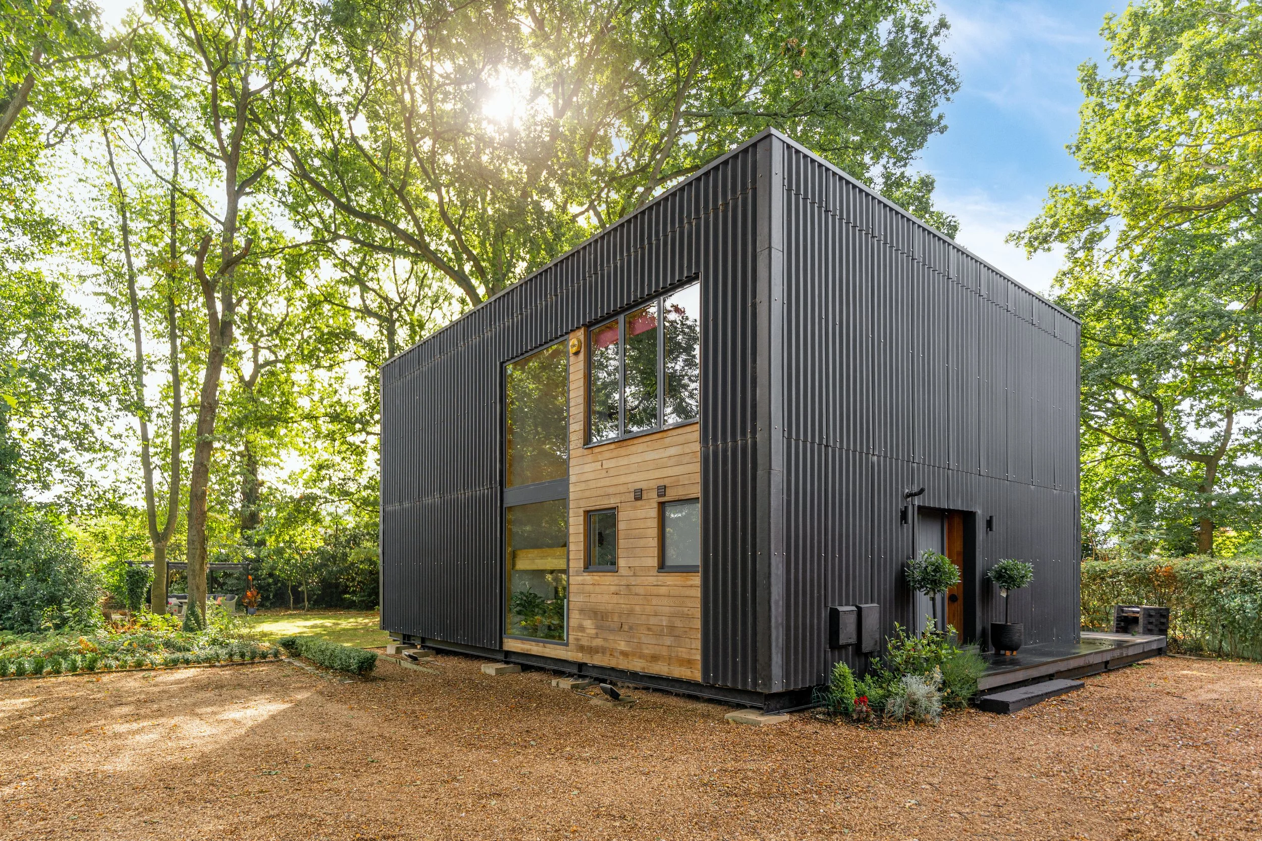 Modern two-story house with black metal siding and wooden accents, surrounded by greenery and tall trees, sunlight filtering through the leaves.