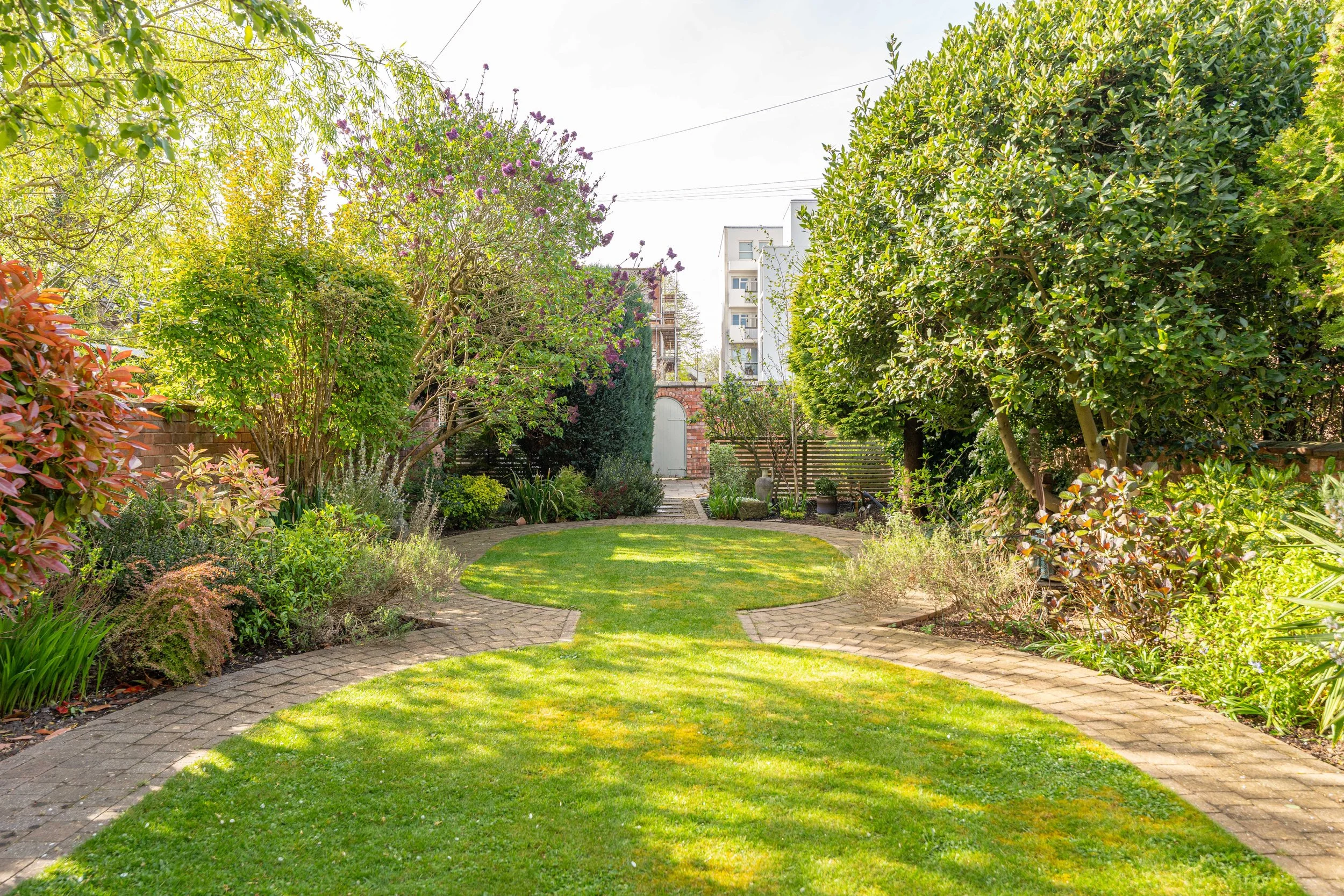 A lush, well-maintained backyard garden with green grass, flowering bushes, and large trees. A paved pathway runs through the garden, leading to a brick archway with a white door in the background. Residential buildings are visible beyond the garden.