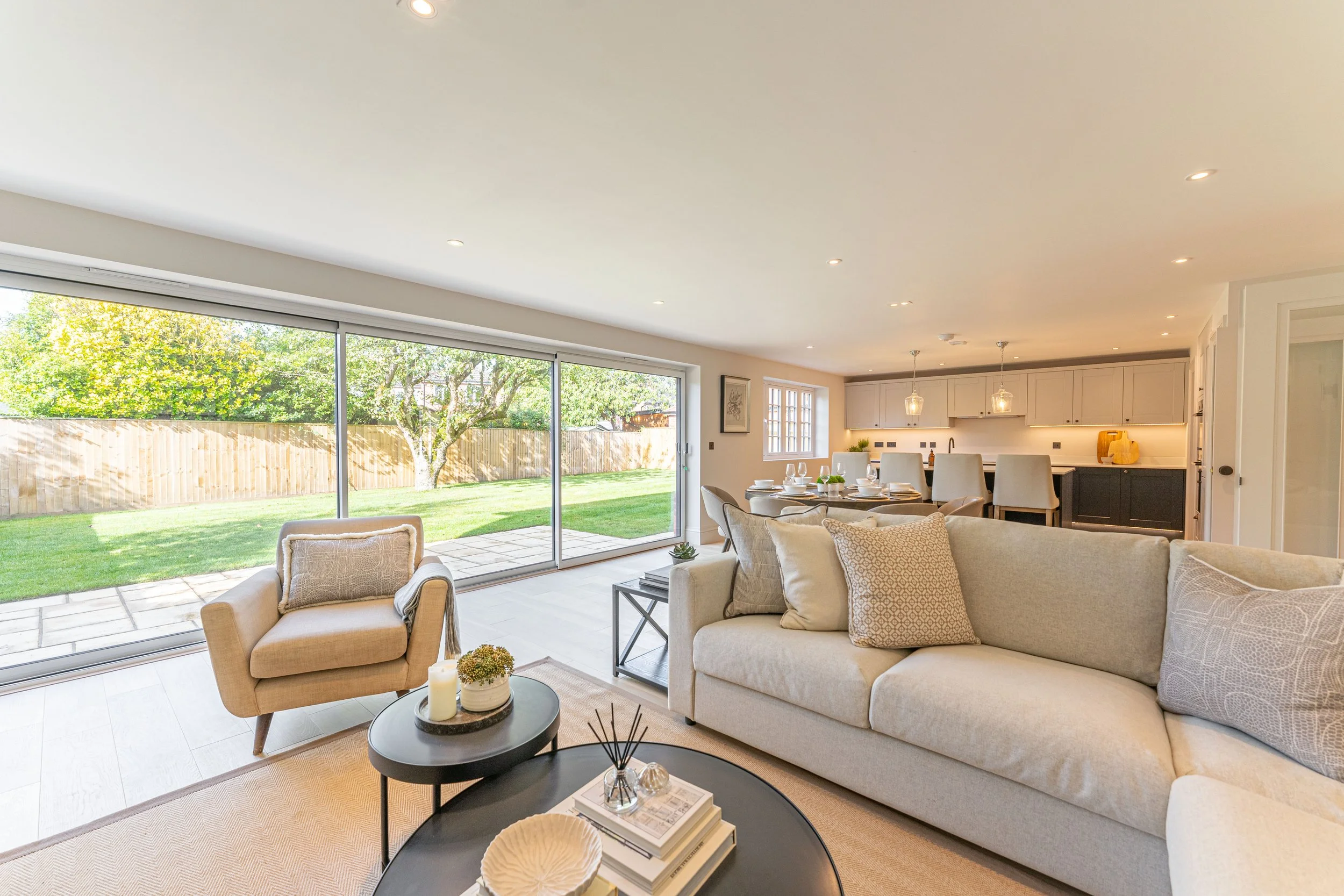 Bright living room with large sliding glass doors leading to a backyard with green grass, trees, and a wooden fence; beige sofa and armchair with decorative pillows; coffee tables with candles, books, and décor; dining area with table and chairs; ope