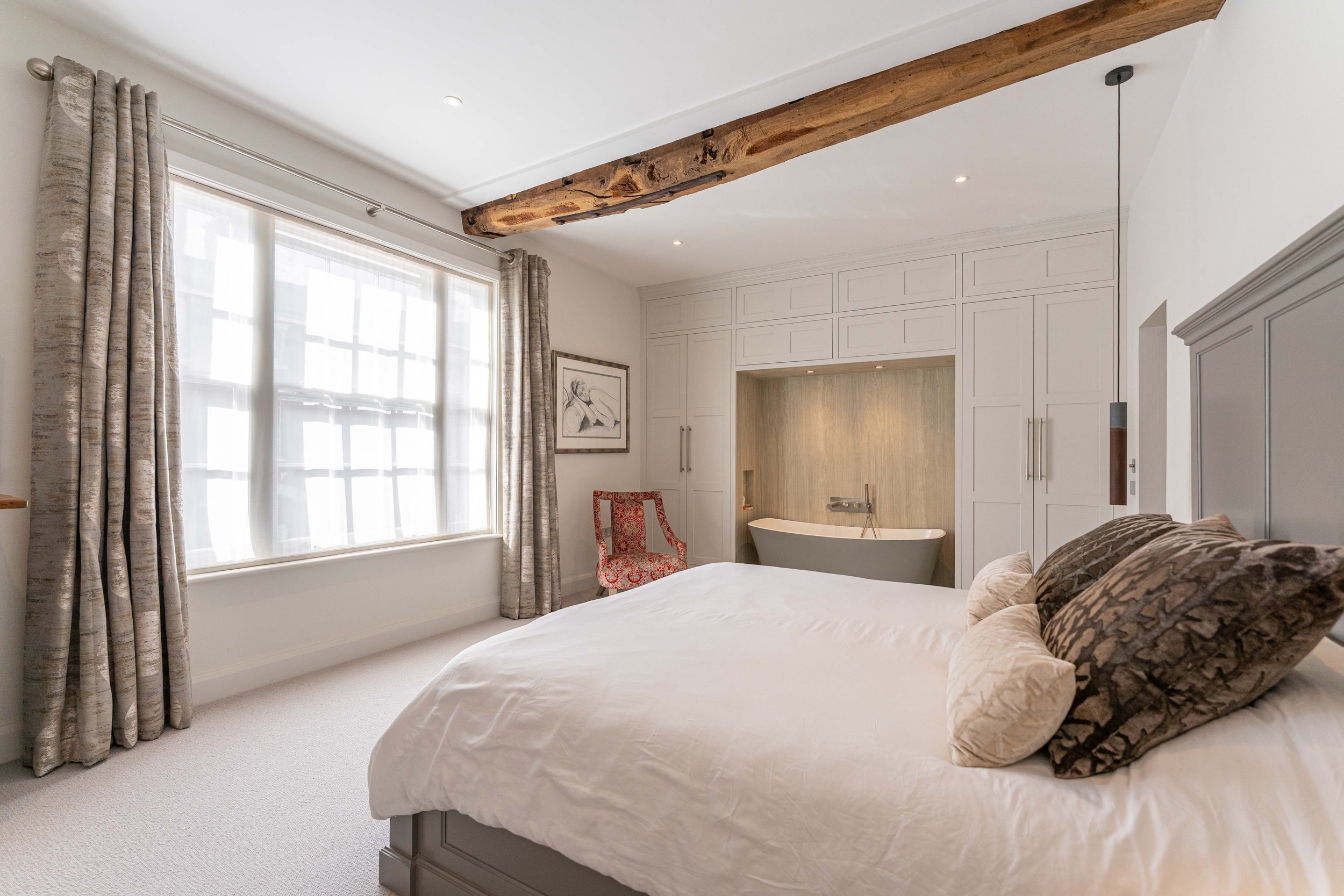 A bedroom with a large window covered by beige curtains, a bed with white bedding and multiple pillows, a red patterned chair, and a bathtub built into a wall with white cabinetry surrounding it.