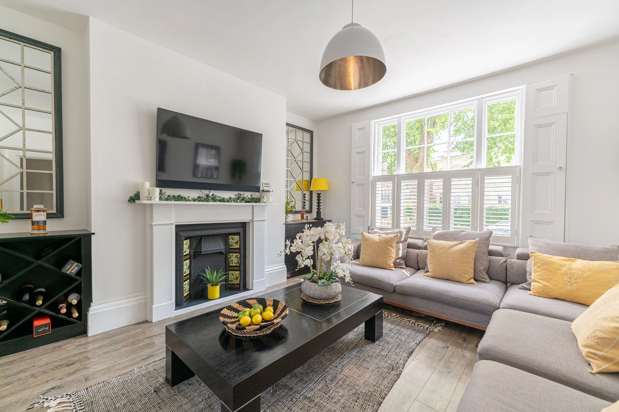 Bright living room with large window, beige sectional sofa with yellow and beige pillows, black coffee table with white orchids, fireplace with candles, yellow lamp, and wall mounted TV.