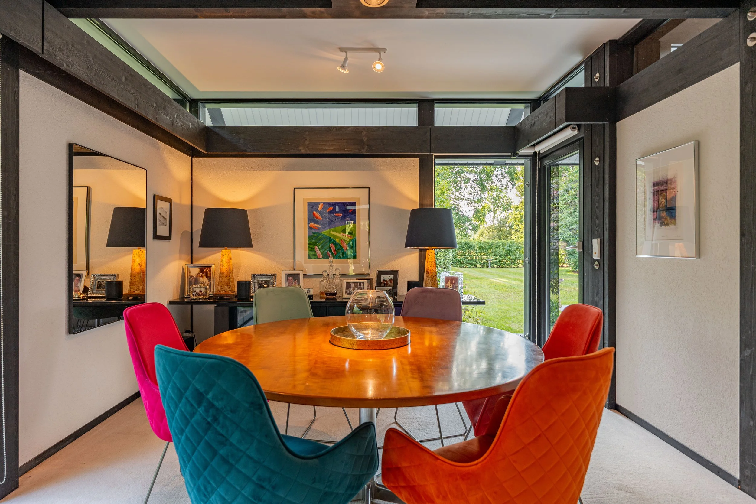 Interior of a modern dining room with a round wooden table surrounded by colorful velvet chairs, large windows with greenery outside, and decorative items and lamps along the back wall.