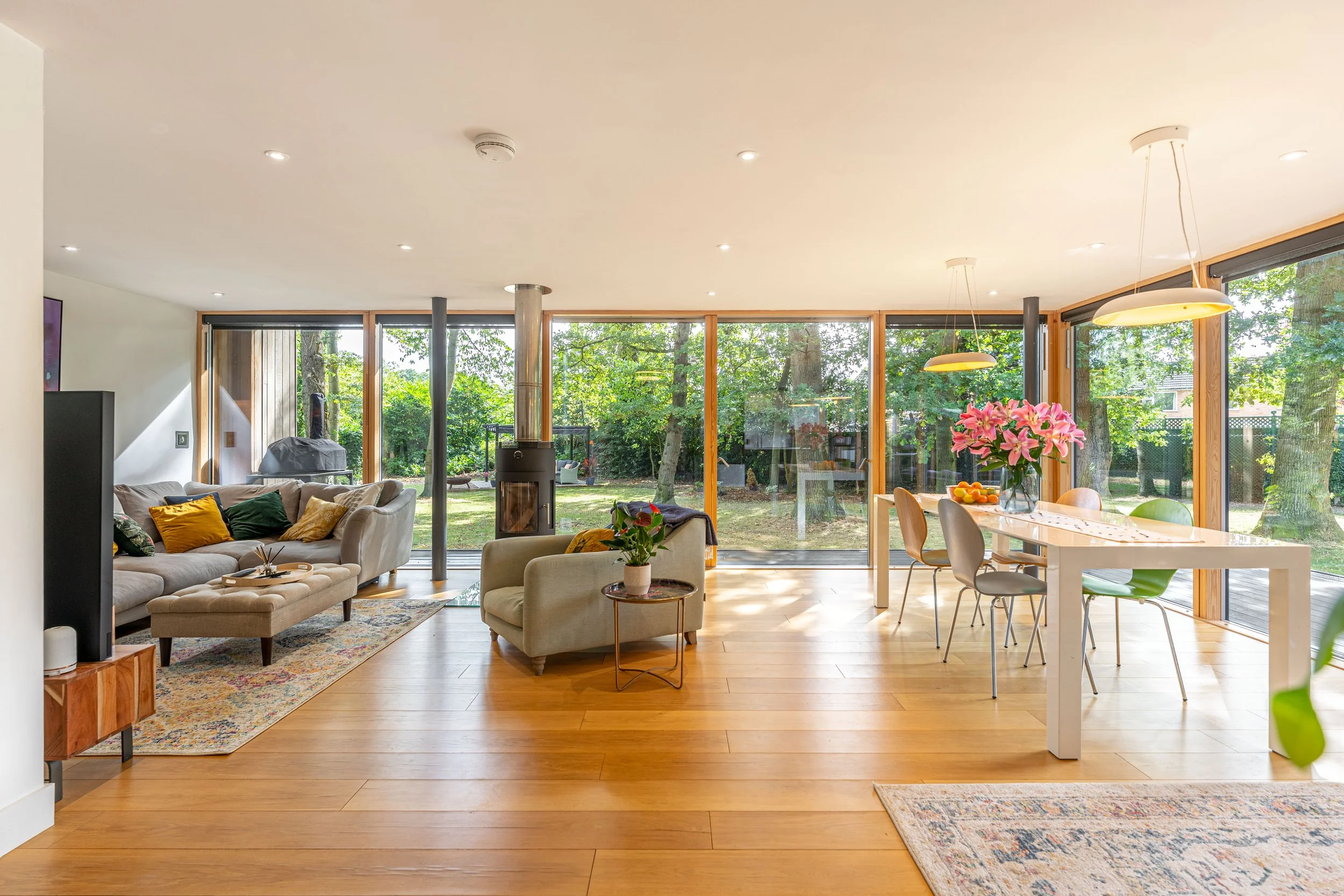 Bright living room and dining area with large windows overlooking a green backyard, a beige sofa with cushions, armchair with a side table, a modern white dining table with chairs, pink flowers in a vase, and a wood-burning stove.