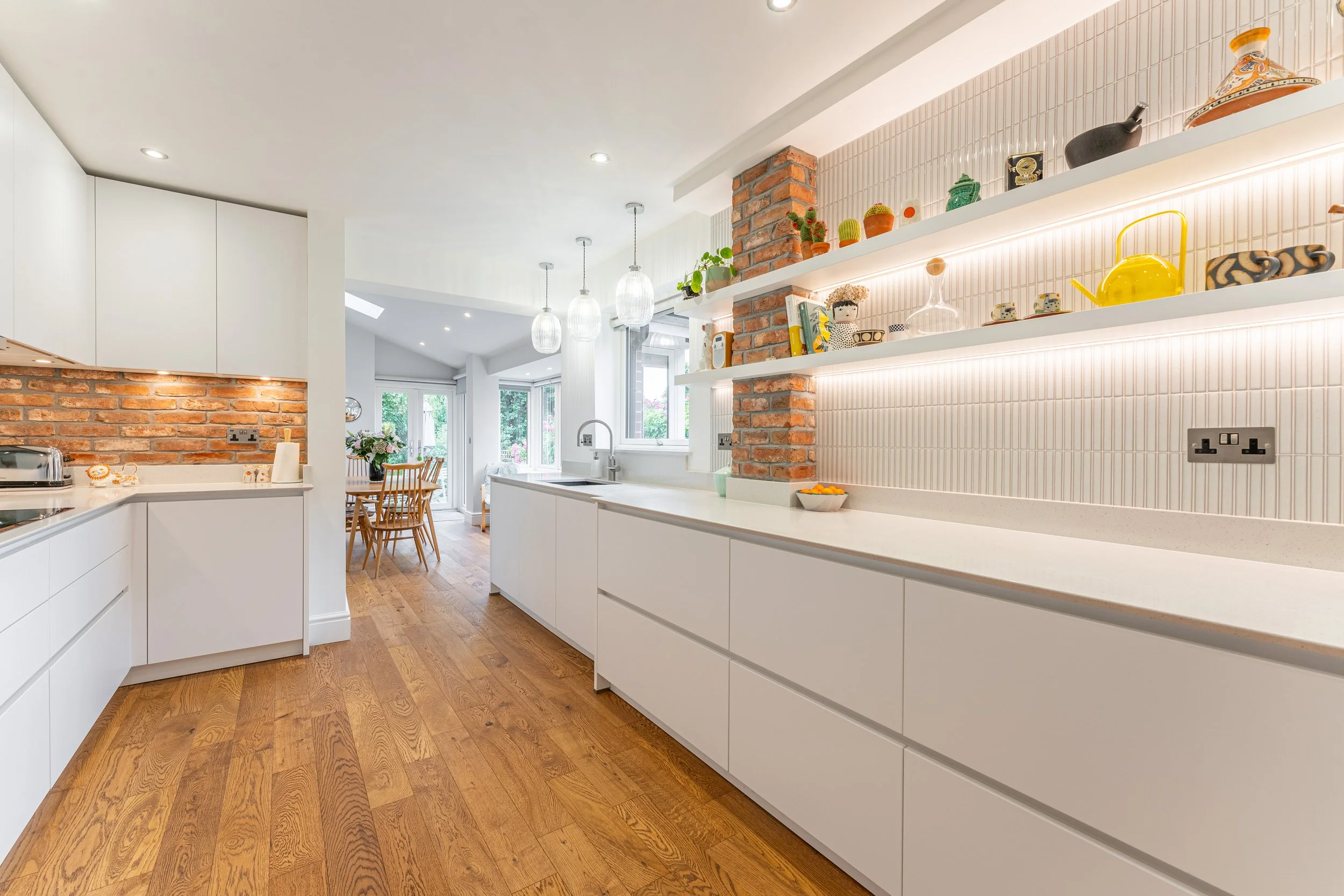 Modern kitchen with white cabinets, brick accents, open shelves, hardwood flooring, and a dining area in the background.