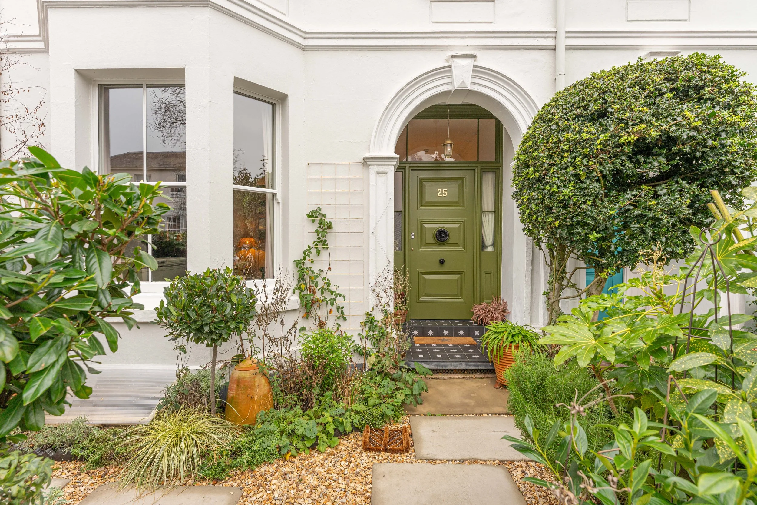 The front entrance of a house with a green door numbered 25, surrounded by various plants, including a large rounded shrub to the right and potted plants, with a gravel pathway and concrete stepping stones leading to the door.